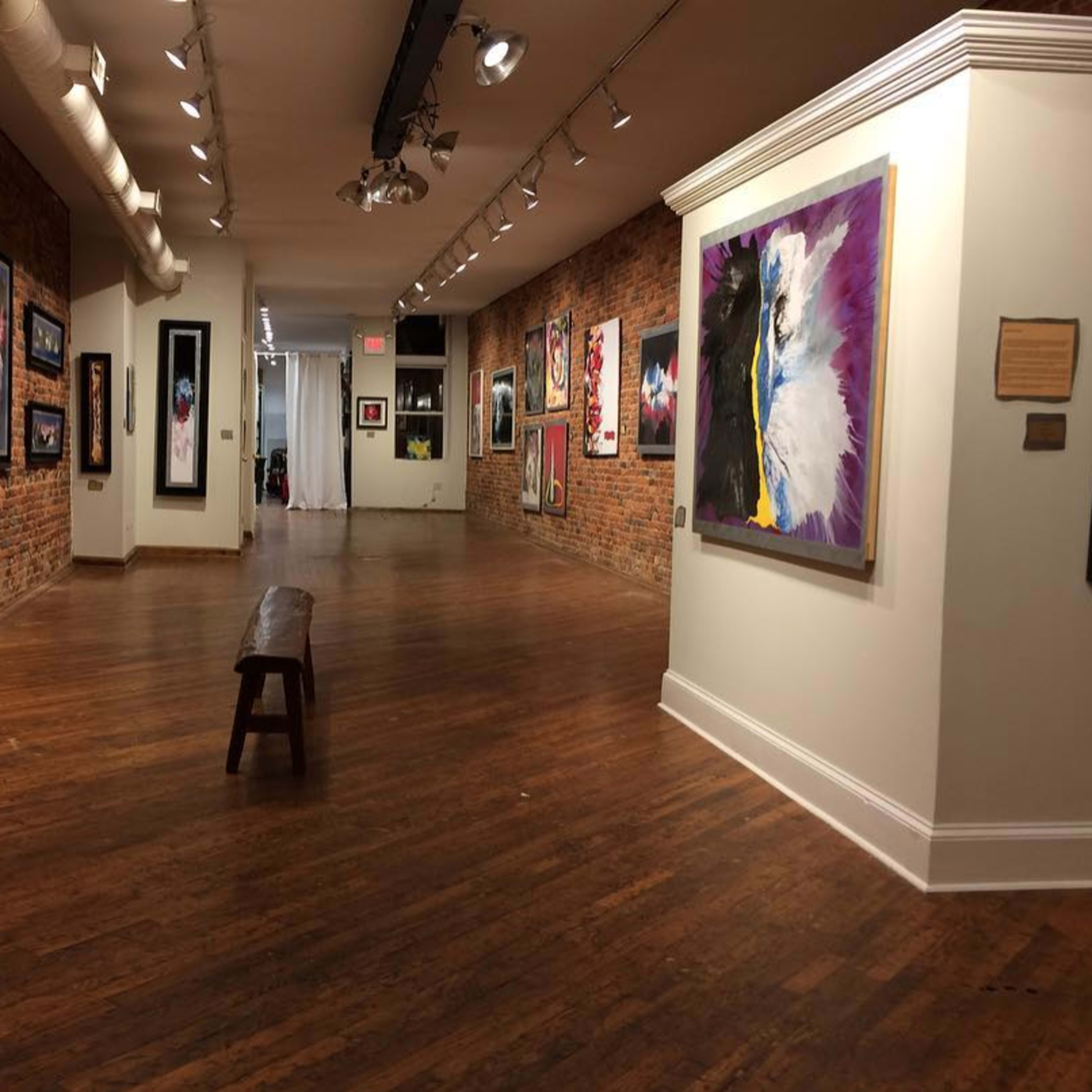 An art gallery with brick walls, wooden flooring, and various paintings displayed on the walls, leading to a white section at the end.