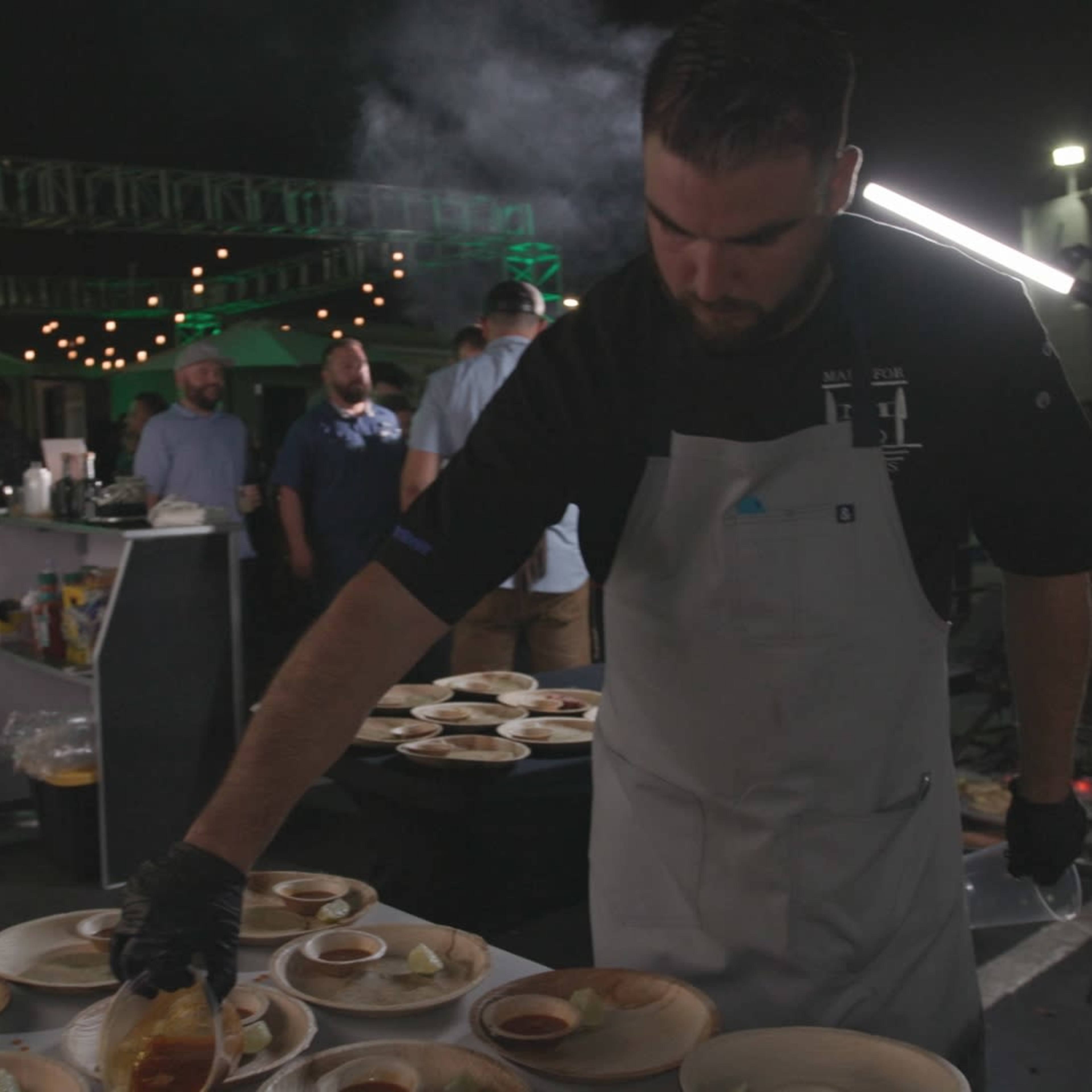 A chef in an apron drizzles sauce over plates of food at a nighttime outdoor event with string lights in the background.