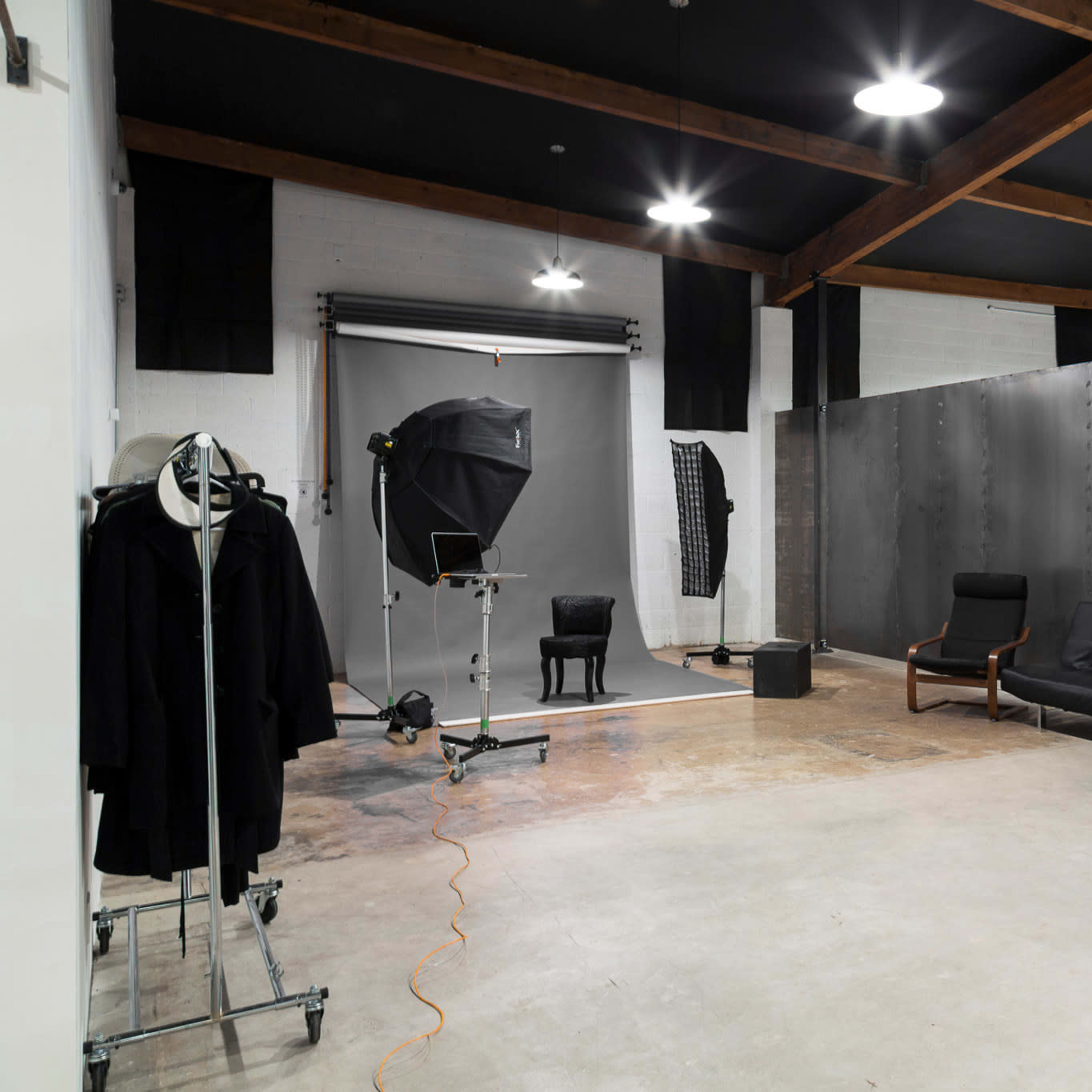 A spacious photography studio featuring a gray backdrop, a lighting setup, a black chair, and a sofa area with minimalist decor.
