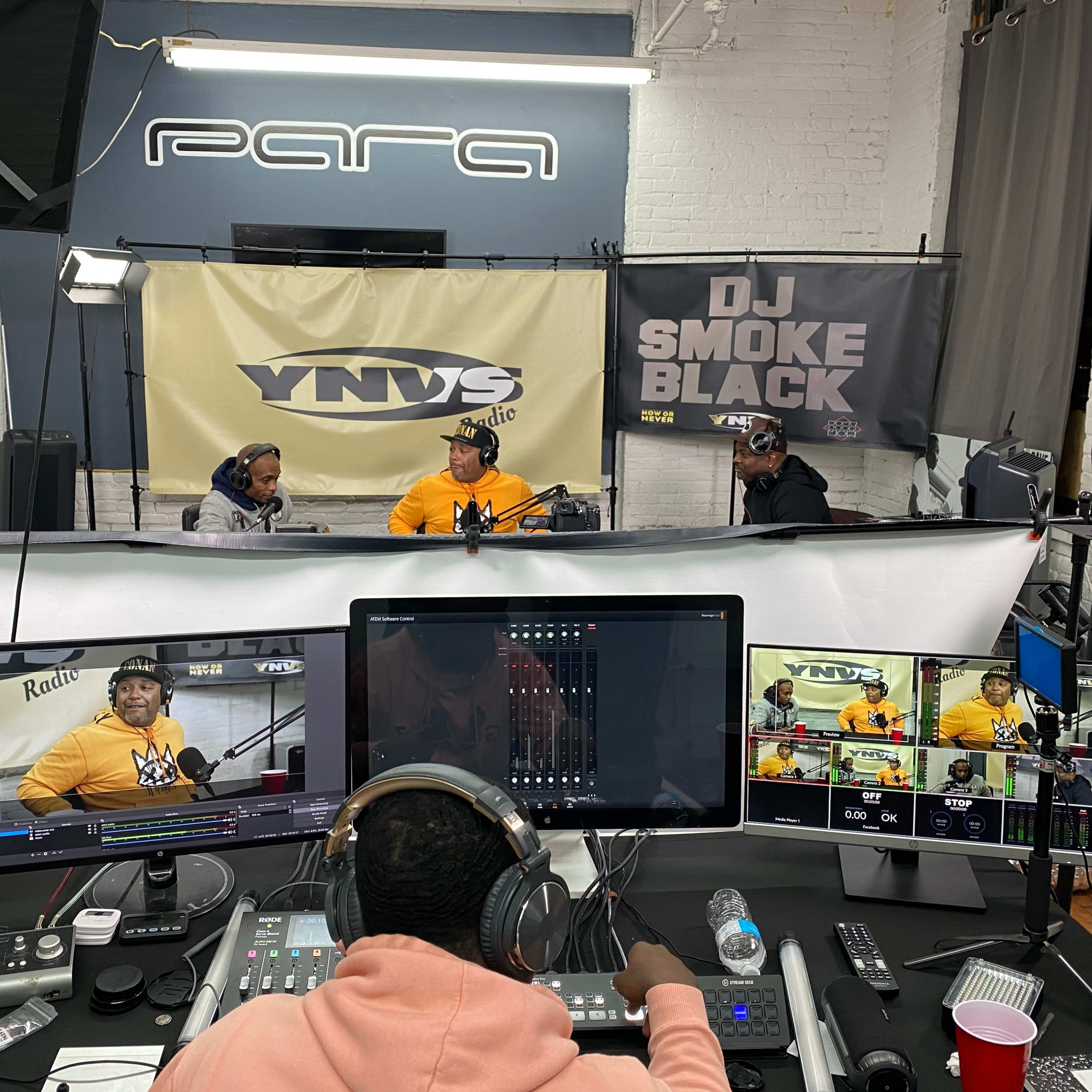 A radio studio setup with three individuals engaged in conversation, surrounded by audio equipment and banners displaying the station's name and DJ's tag.