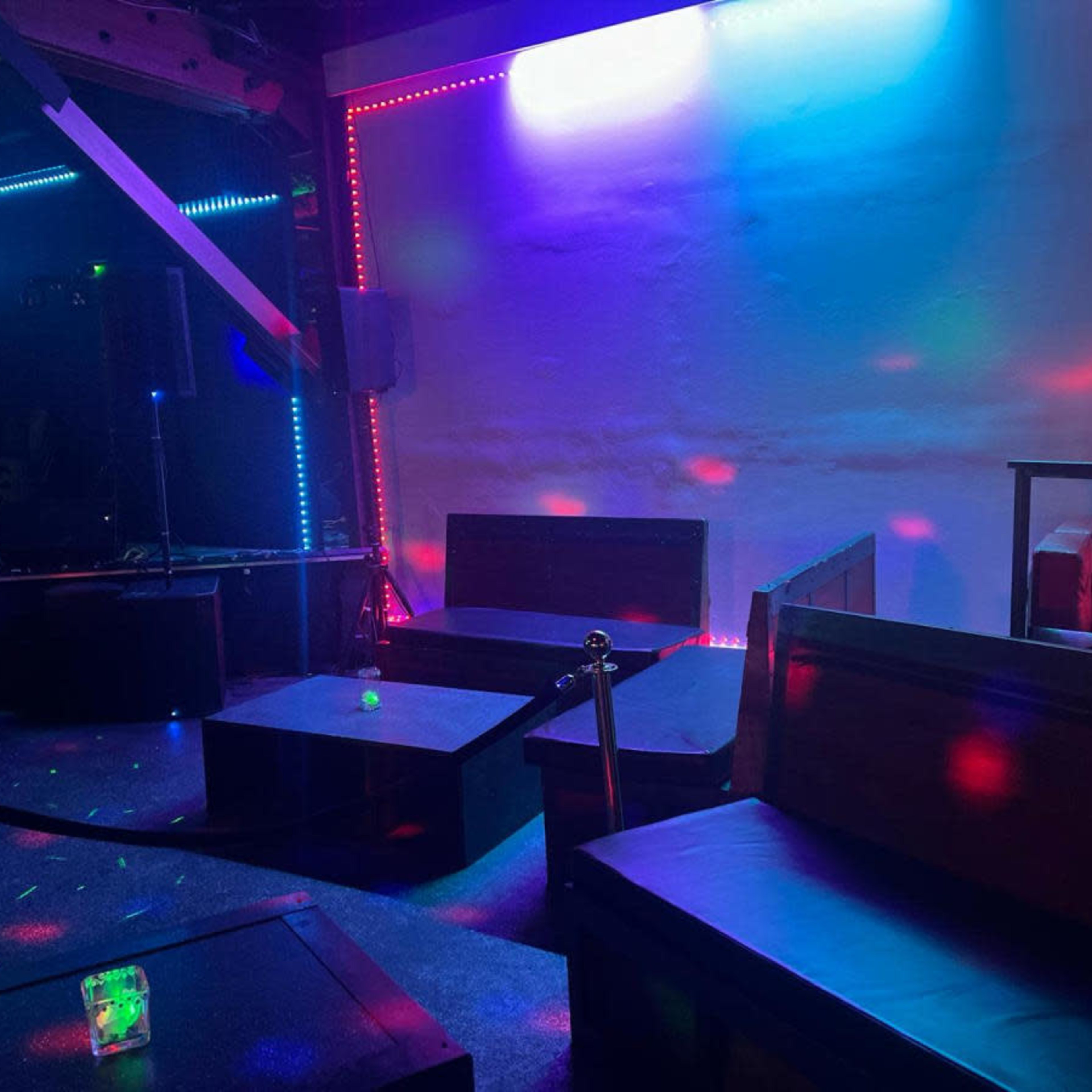 A dimly lit lounge area features several black couches and tables, illuminated by colorful lights along the walls.