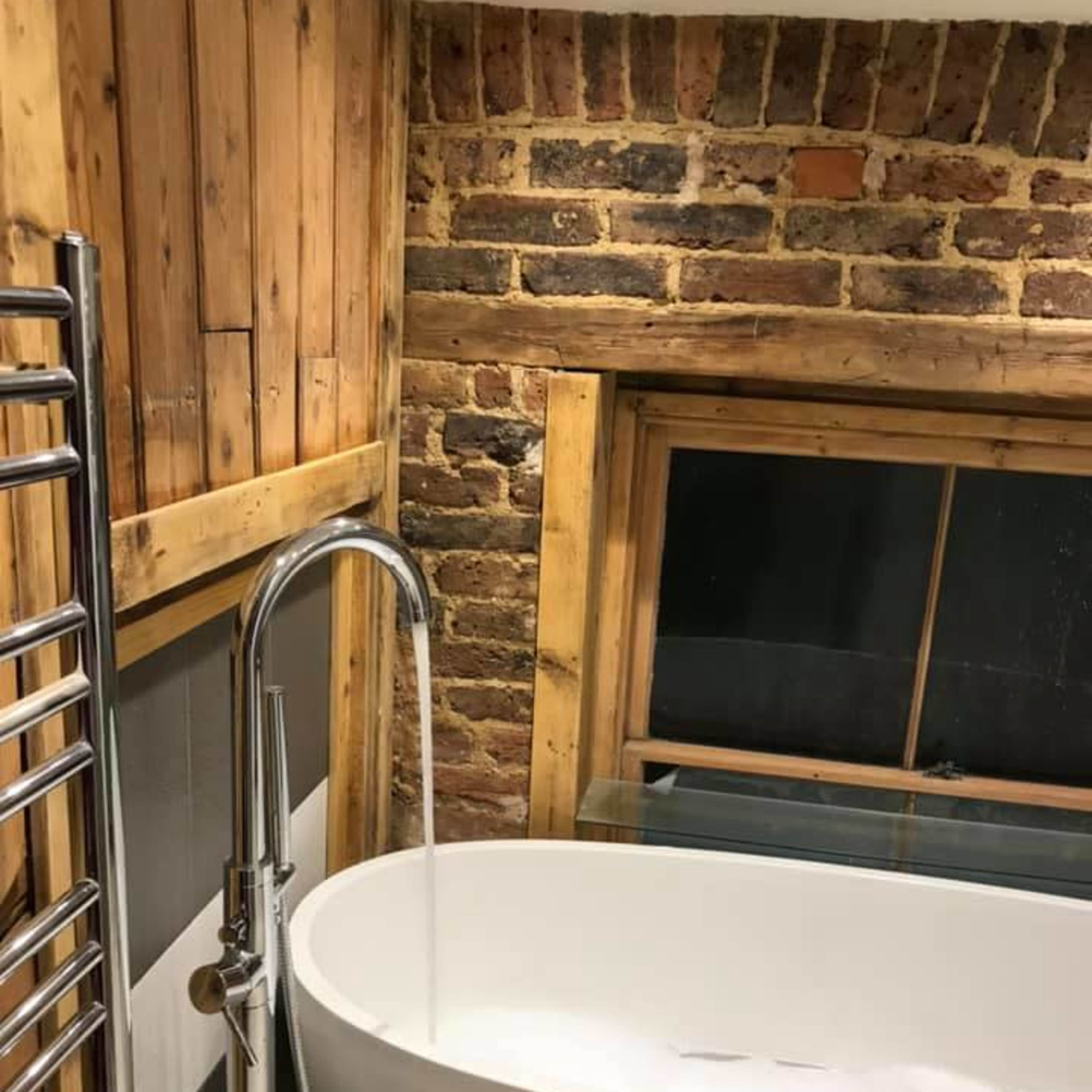 A modern bathroom featuring a freestanding white bathtub next to a wooden-paneled wall and exposed brick, with water flowing from a sleek faucet.