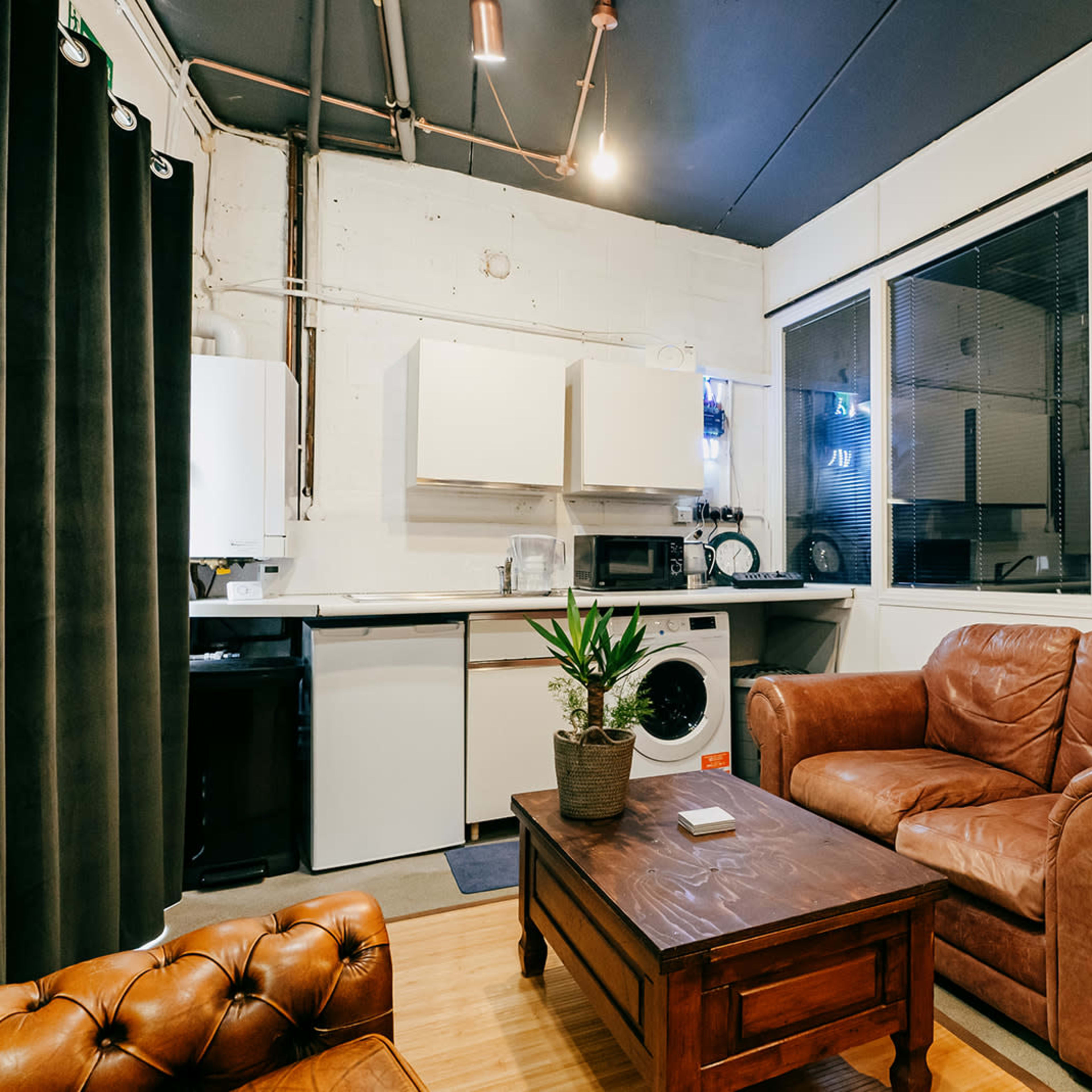 A cozy living area with a brown leather sofa, a wooden coffee table, and a kitchenette featuring white cabinets and various appliances.