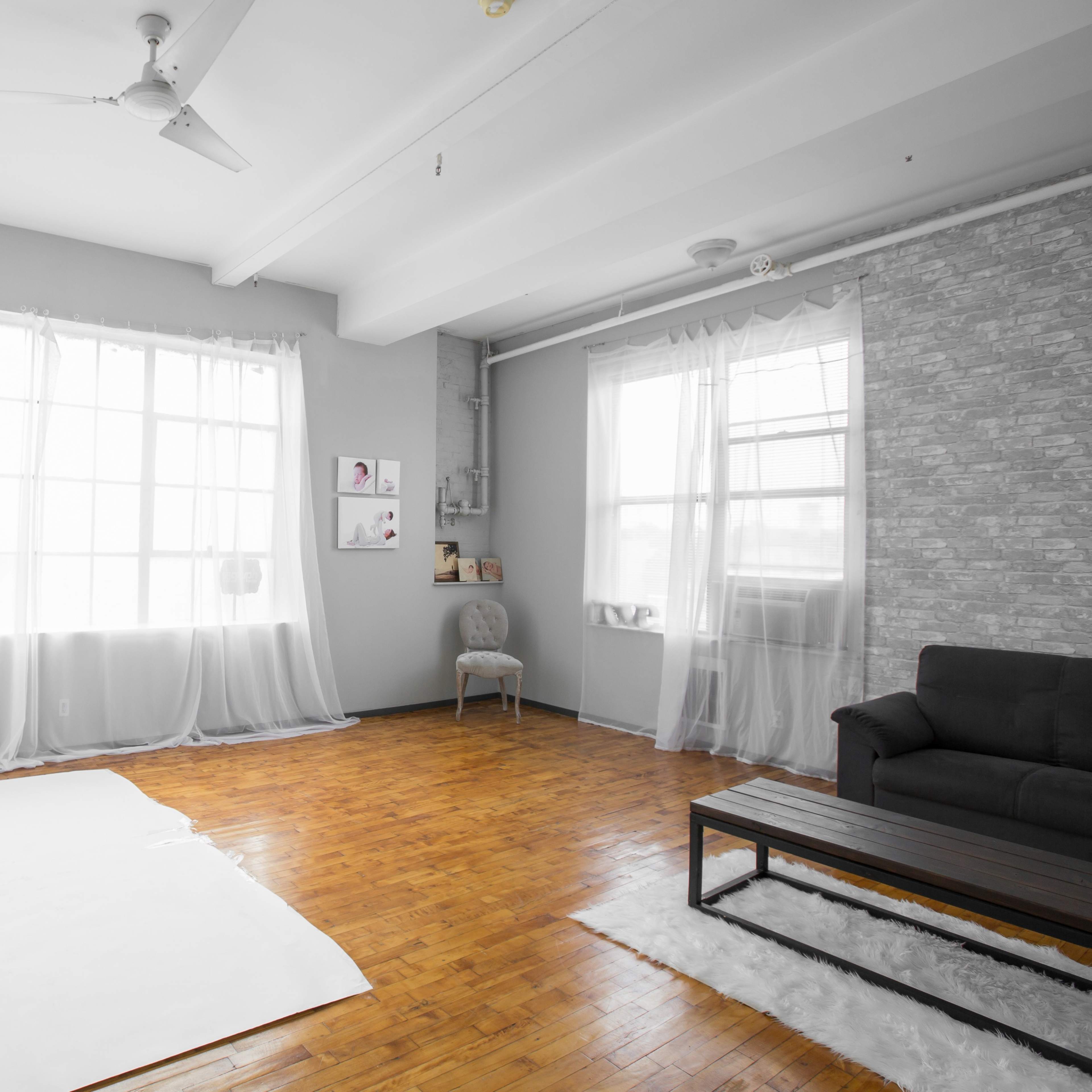 A simple, bright room with wooden flooring, large windows covered by sheer curtains, and minimal furnishings including a dark sofa and a coffee table.