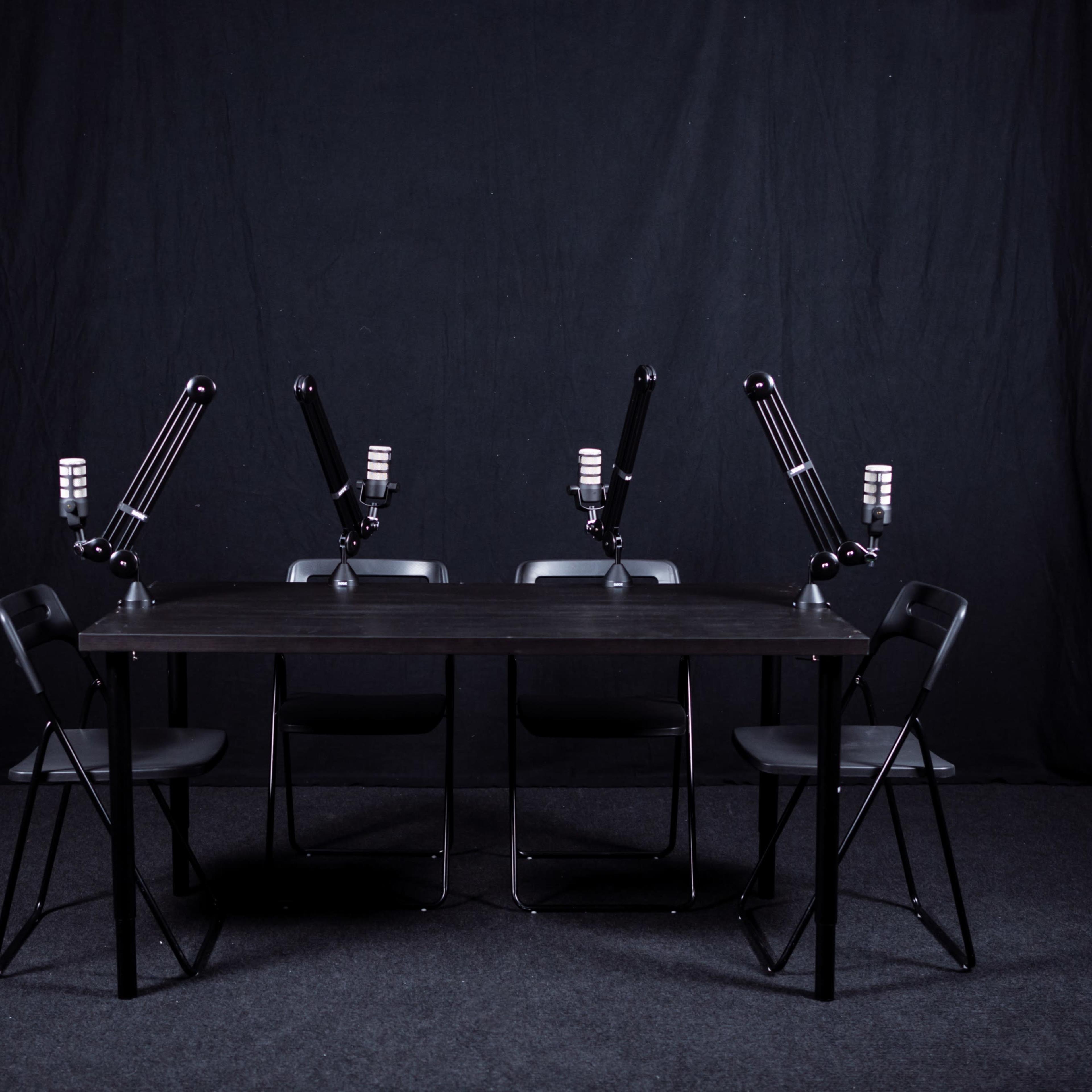 A black table with four chairs and mounted microphones is set against a dark backdrop.