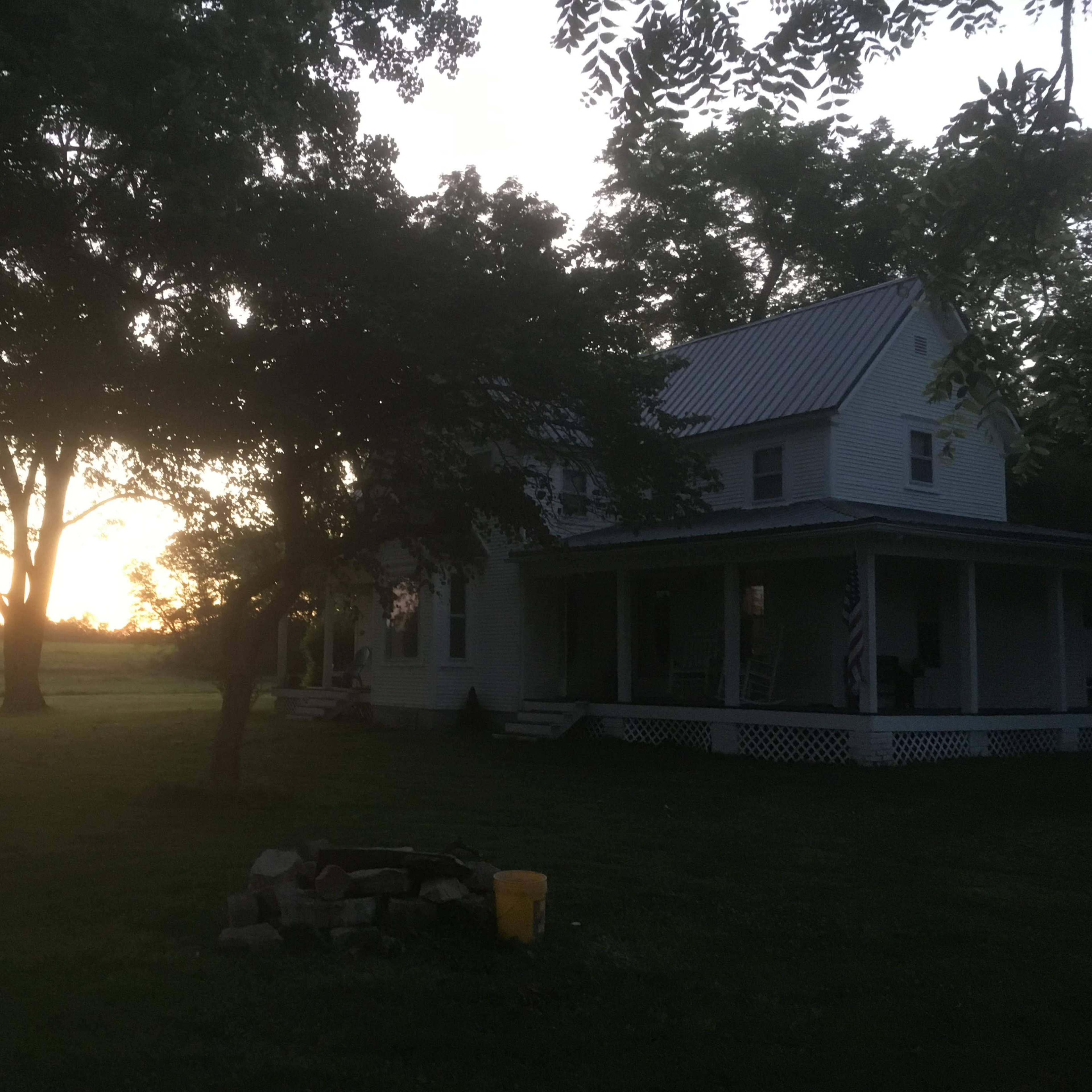 A farmhouse with a porch sits under trees at twilight, with a sunset visible in the background.