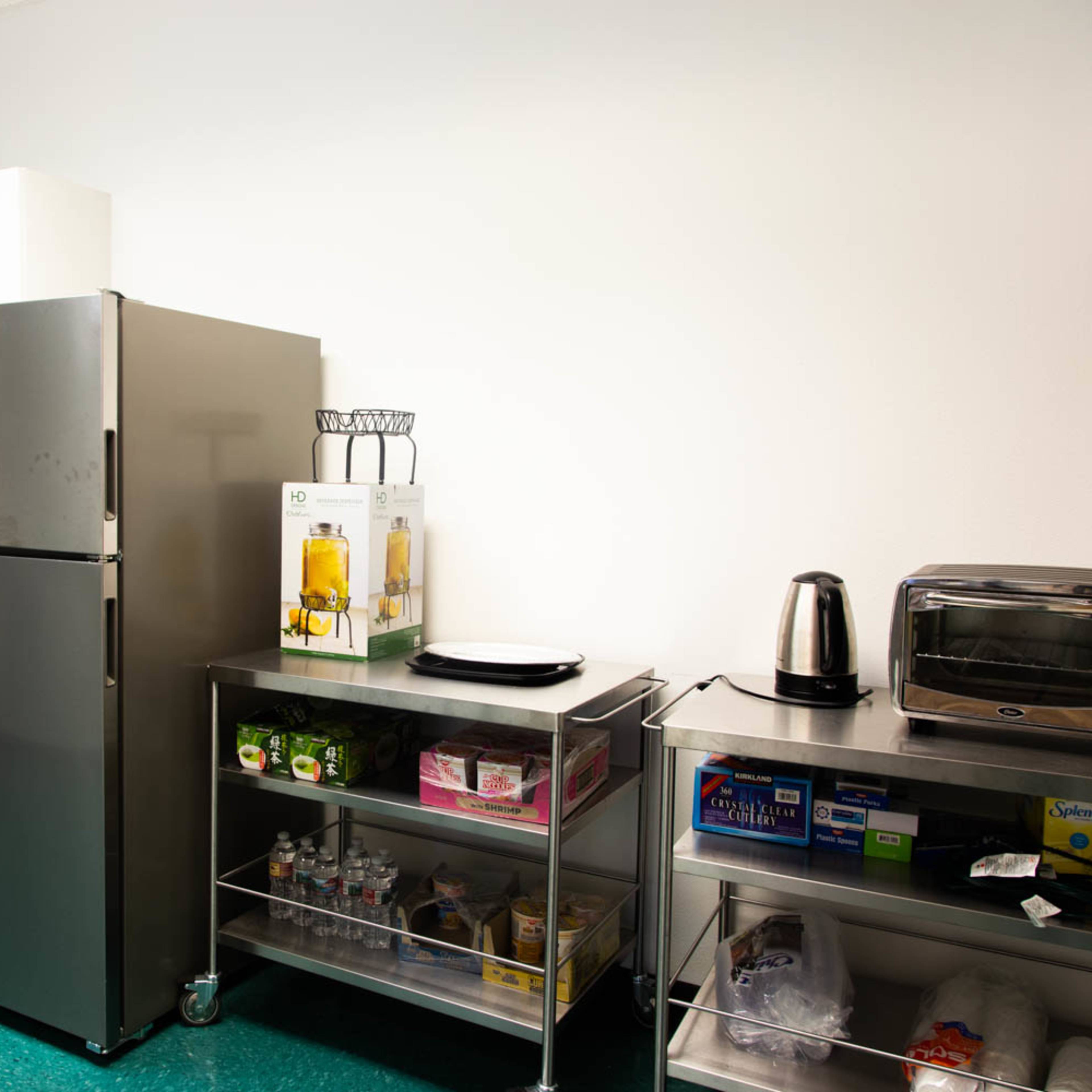 A kitchen area with a stainless steel refrigerator, a shelf filled with drinks and snacks, a toaster oven, and a kettle on a countertop.