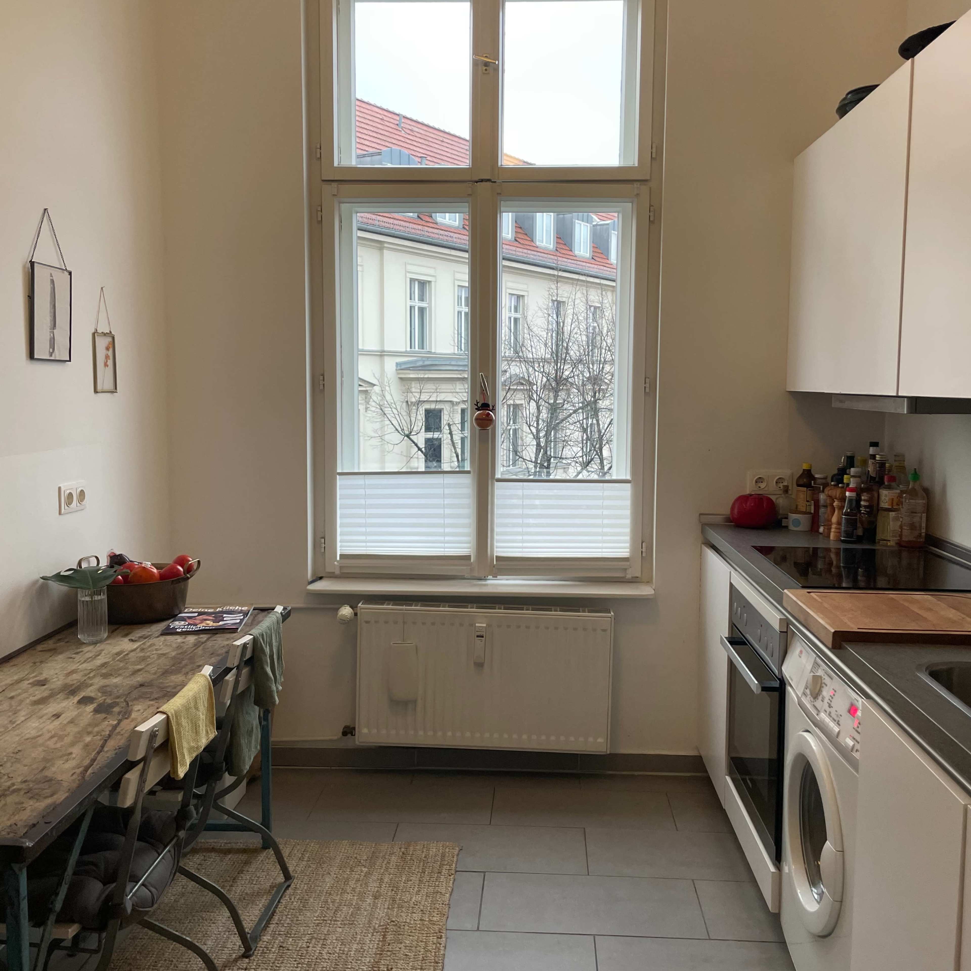 A small kitchen features a wooden table with two chairs, a washing machine, and a large window overlooking a building exterior.