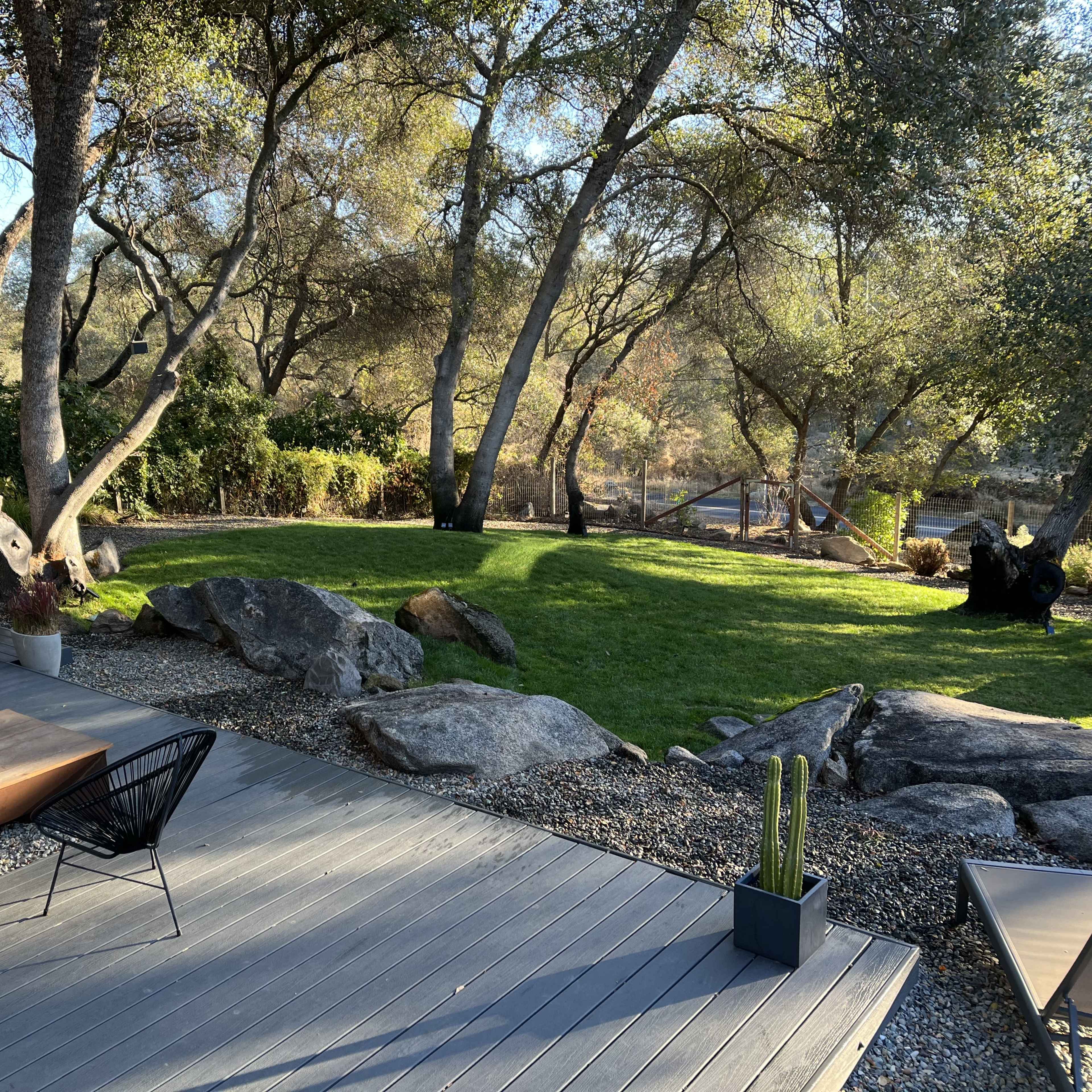 A wooden deck features seating and decorative plants, surrounded by trees and rocks, with a grassy area in the background.