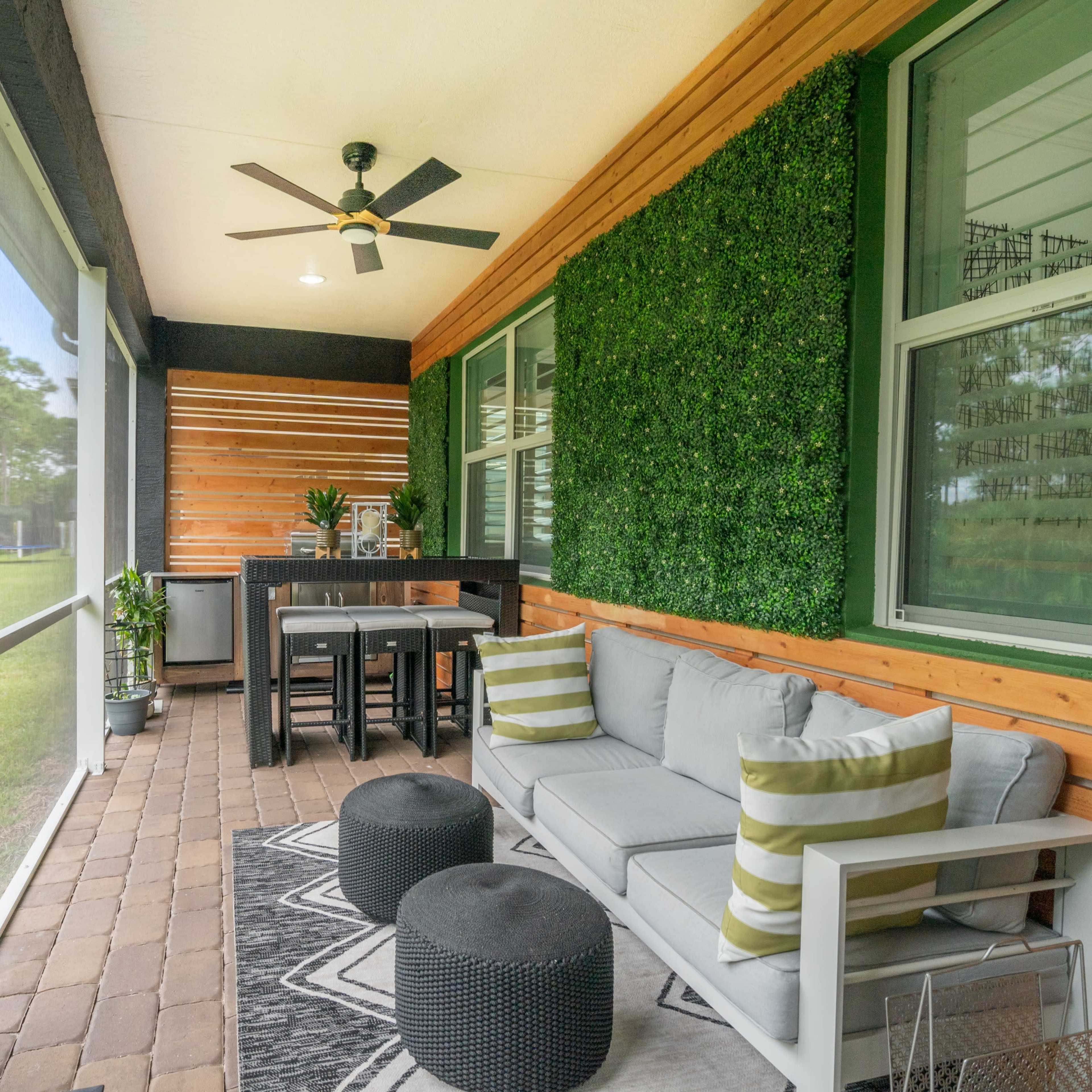 A screened porch featuring a seating area with a couch, two ottomans, and a small dining table, surrounded by greenery and wooden paneling.