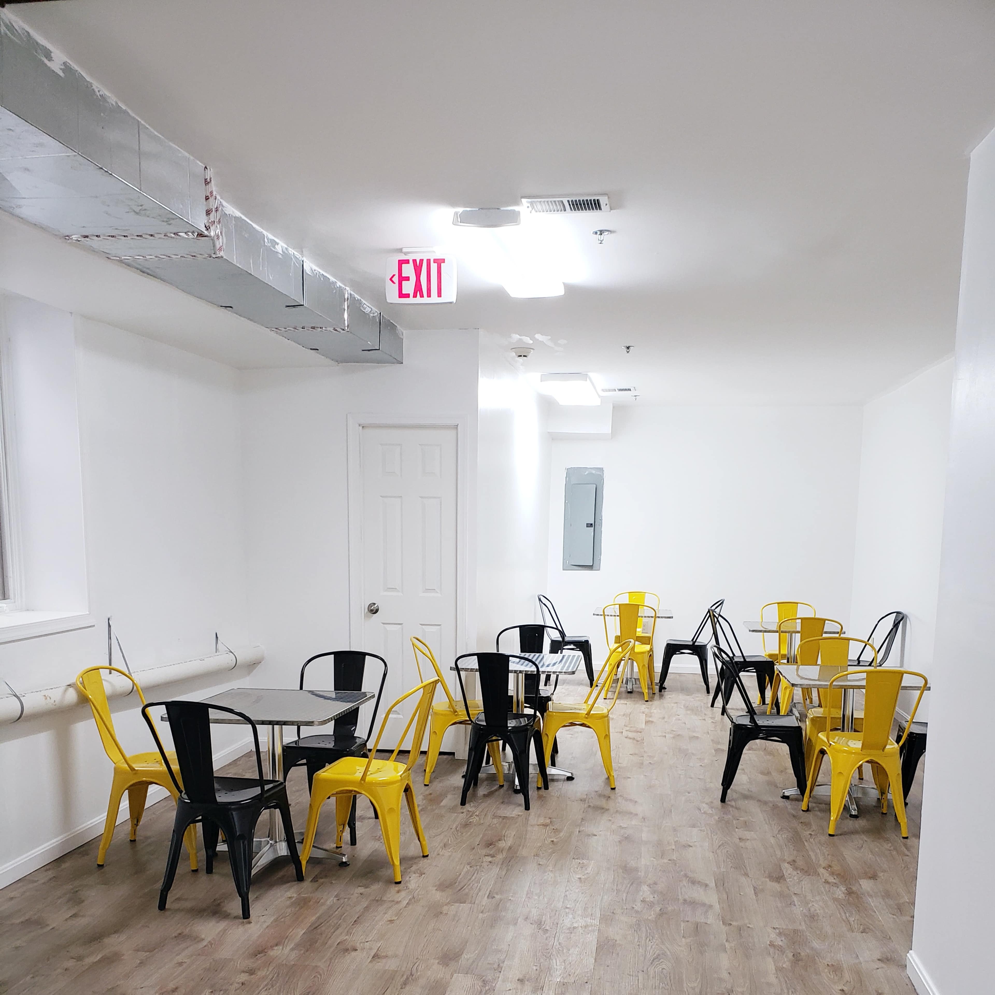 A spacious, well-lit room with wooden flooring, featuring several metal tables with black and yellow chairs arranged throughout.