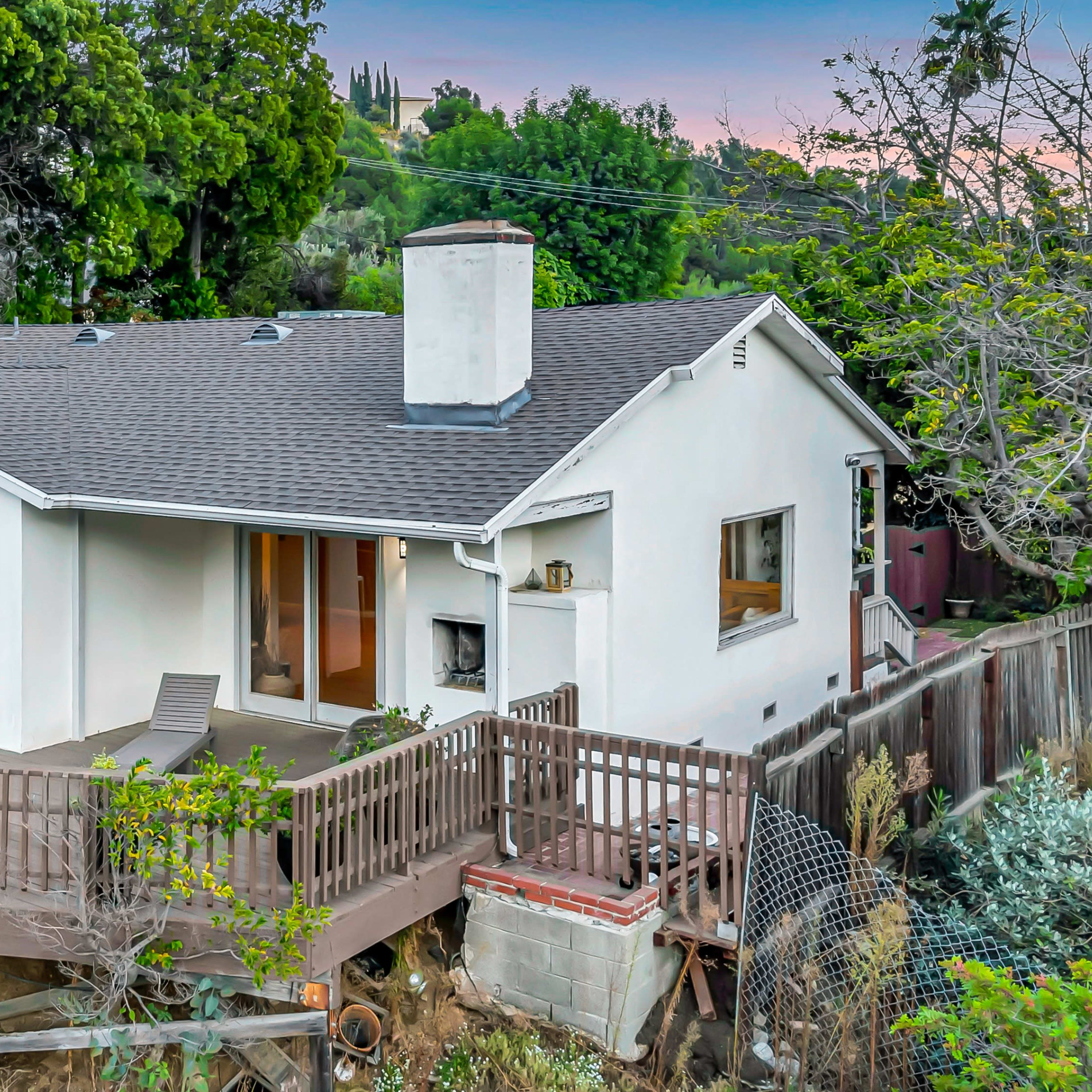 A single-story house with a wooden deck, surrounded by trees and located on a slope.