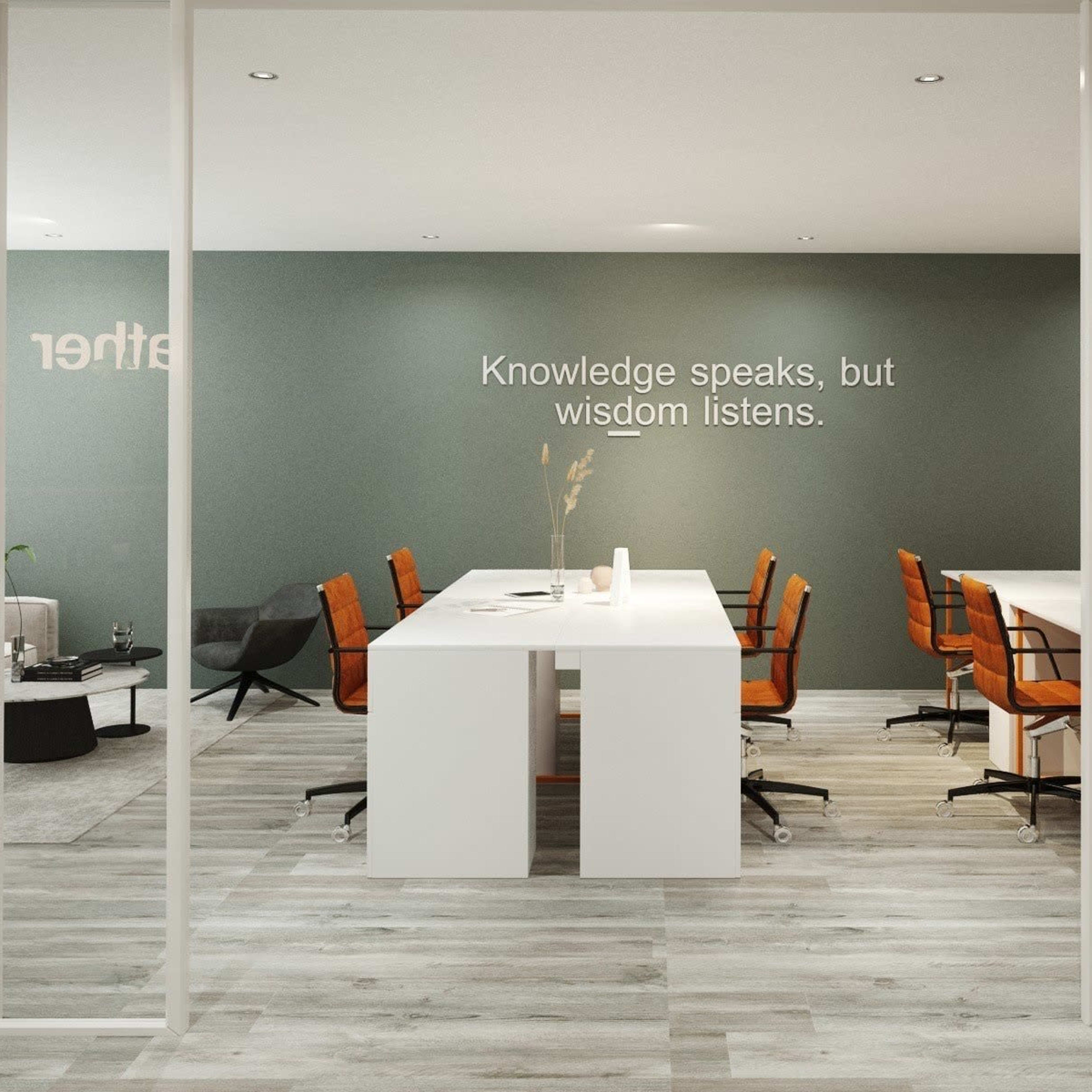 A modern office space featuring a row of white work desks with orange office chairs, a sofa area, and a wall with a quote about knowledge and wisdom.