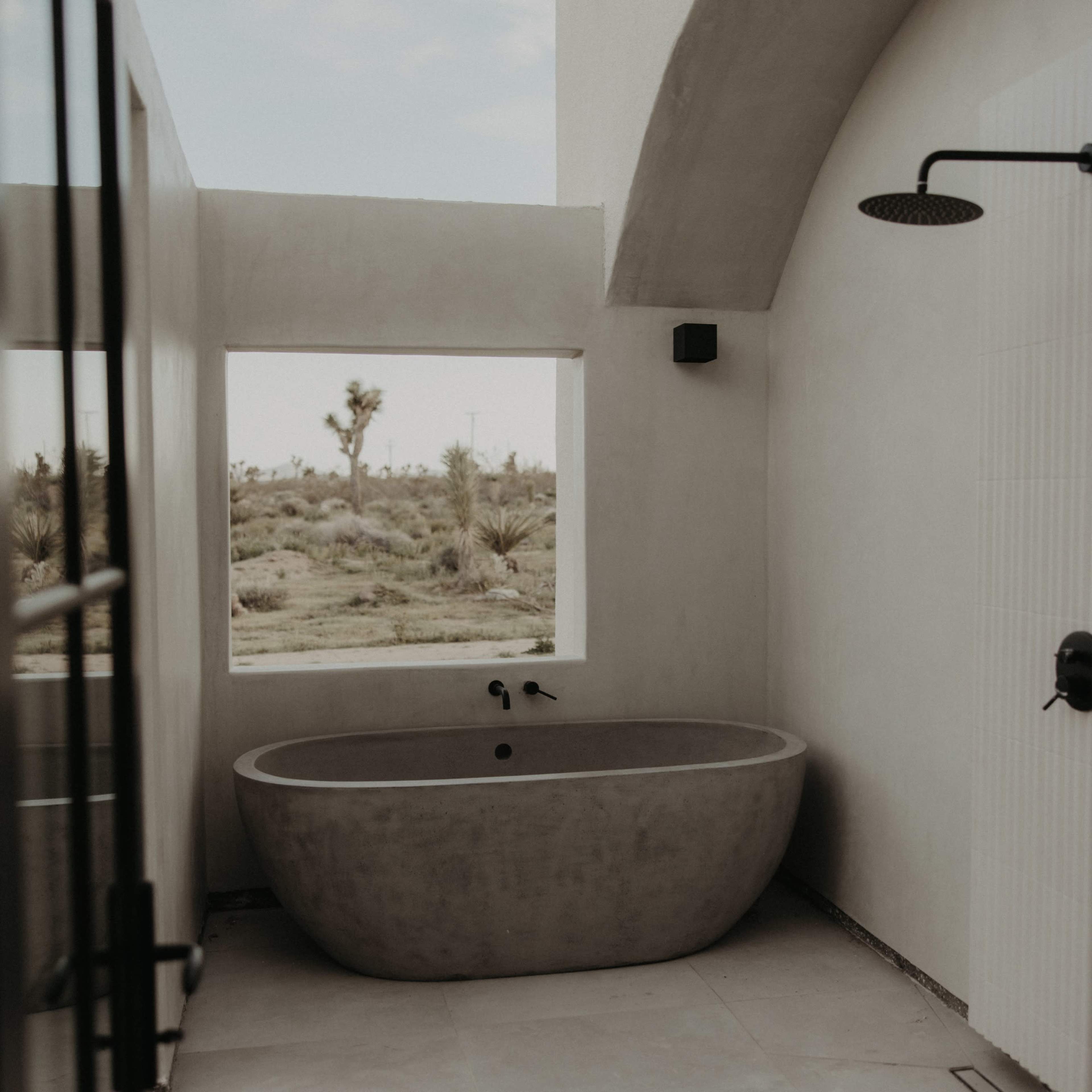 A minimalist bathroom features a concrete bathtub positioned near a large window with a view of desert vegetation.