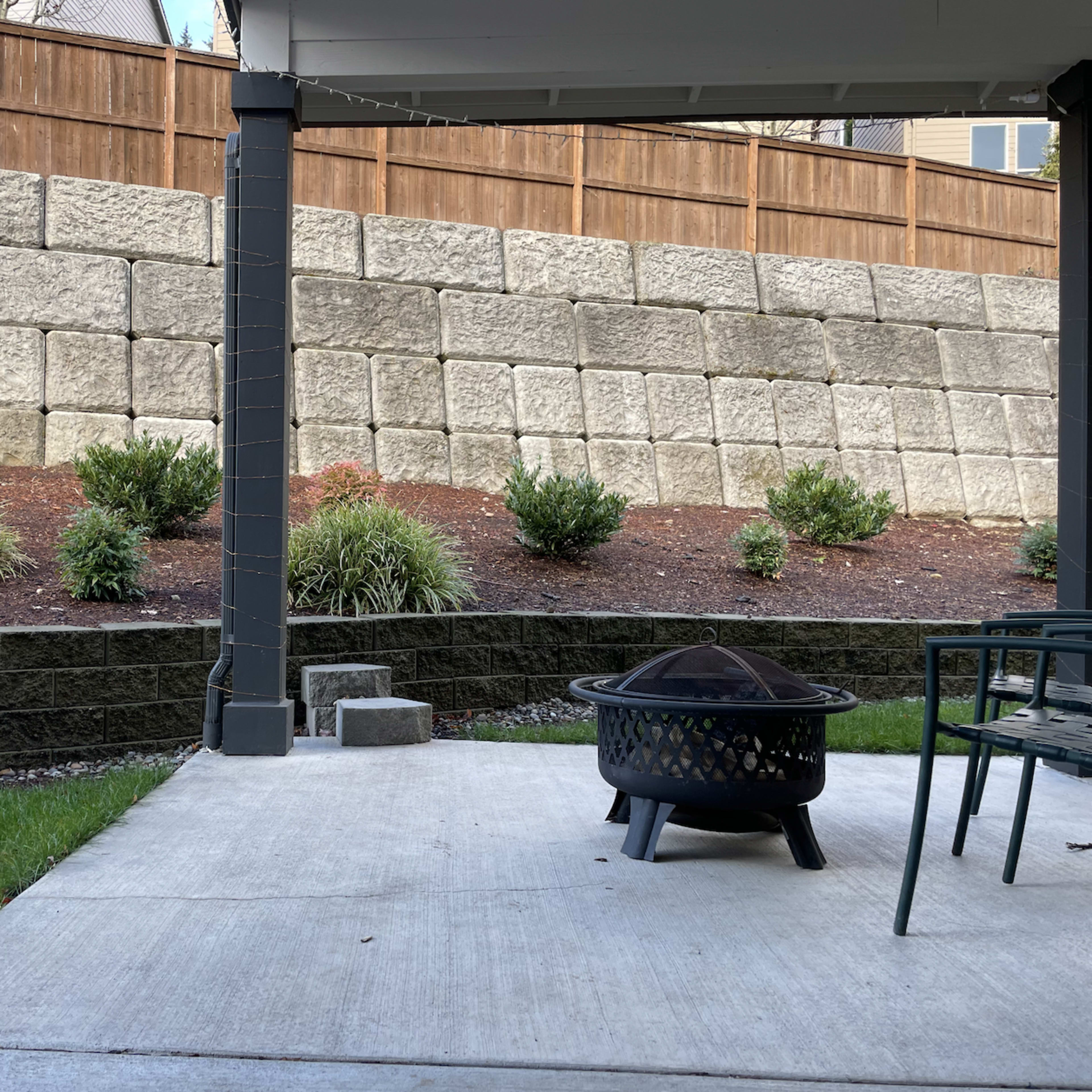 A patio area with a fire pit, surrounded by a stone wall and landscaped with small shrubs.