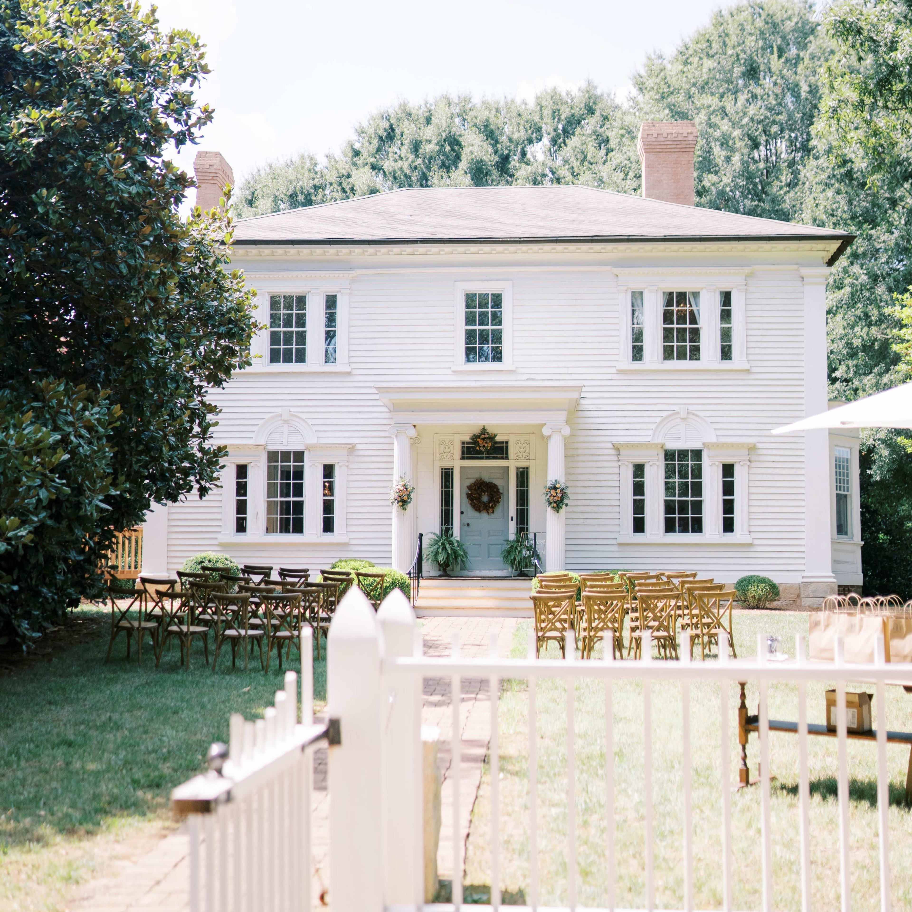 A white, two-story house with a central entrance is surrounded by chairs arranged for an event in the front yard.