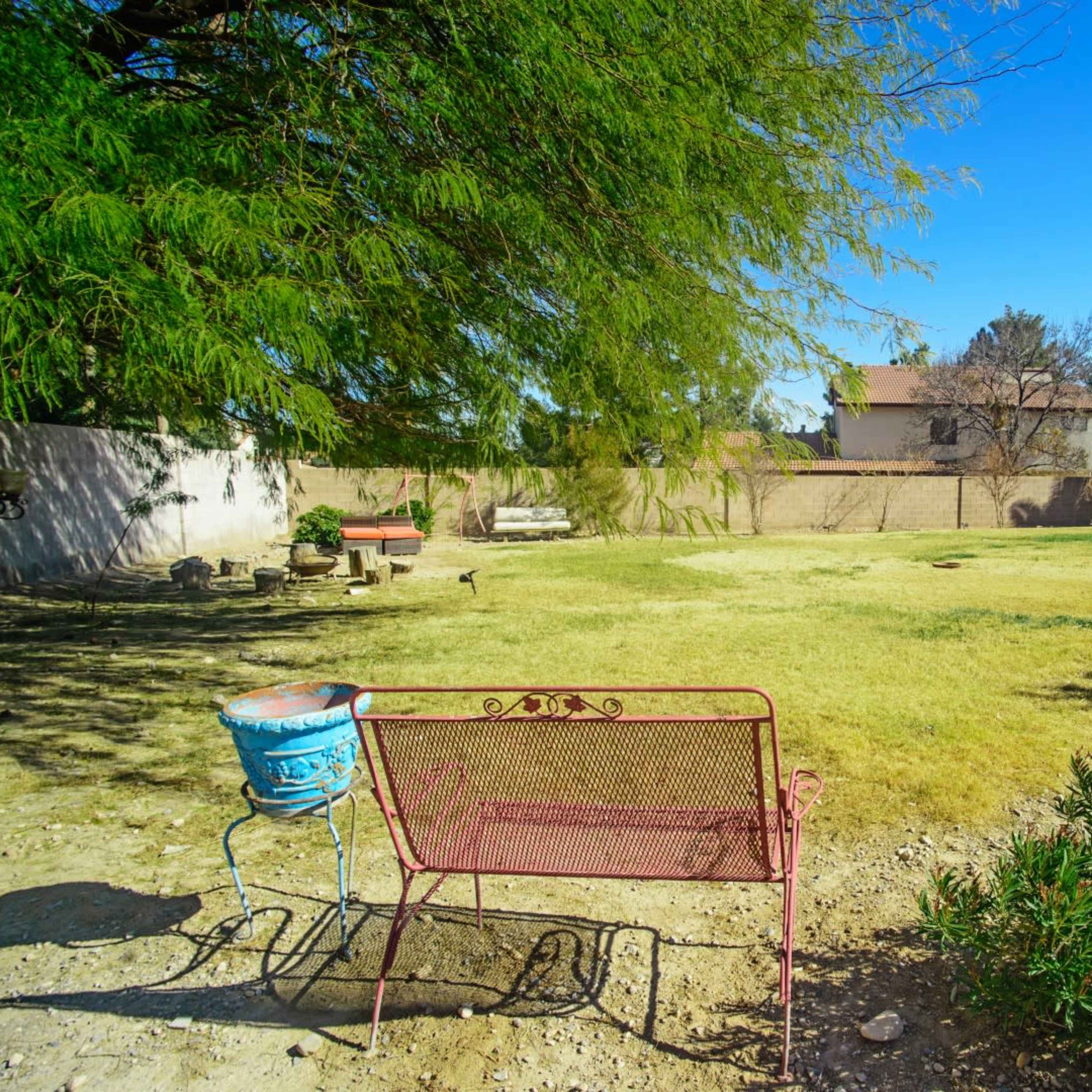 A metal bench sits on the bare ground of a grassy backyard, surrounded by trees and a few structures.