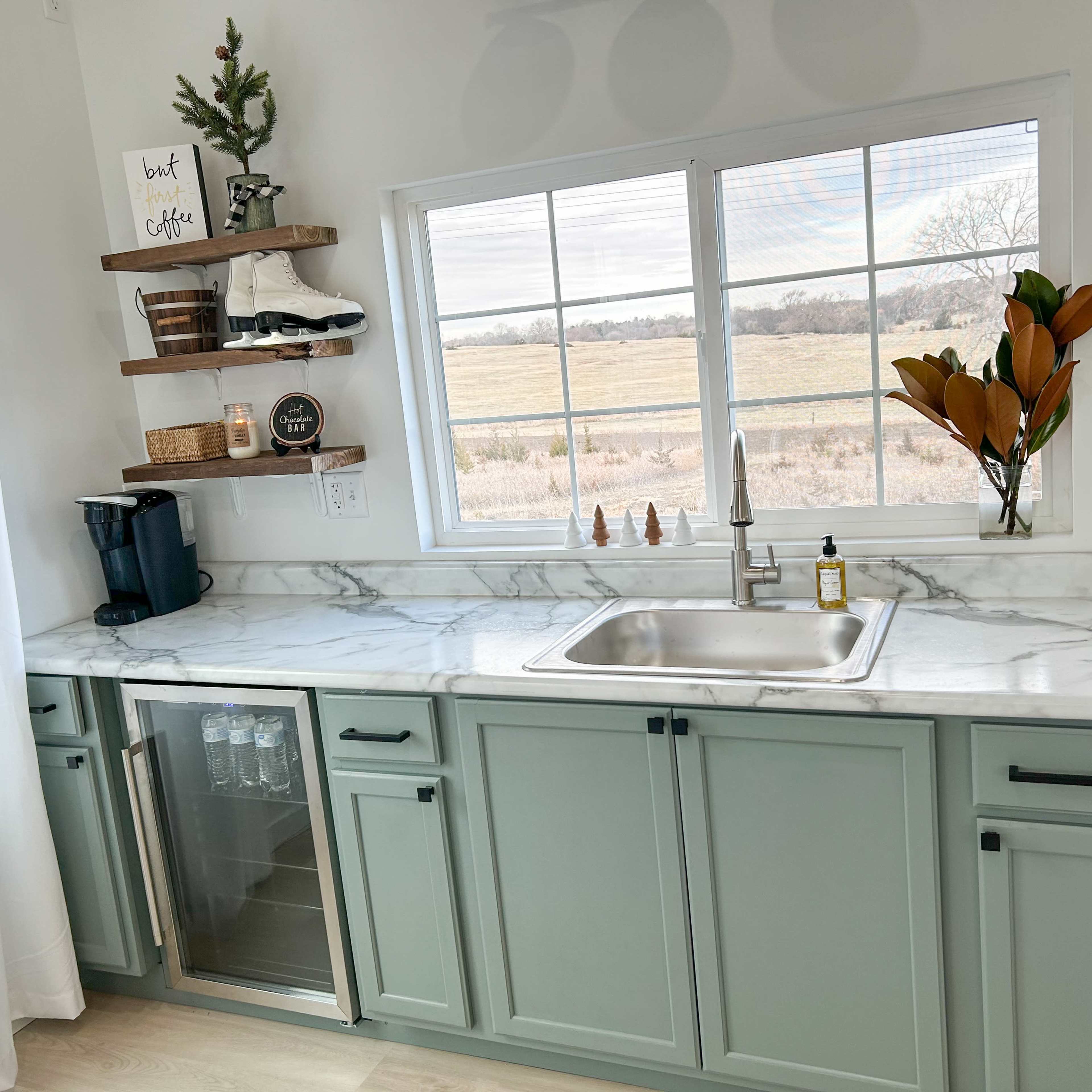 A well-organized kitchen counter features a marble surface, a sink, a mini-fridge, and shelves with decorative items against a window overlooking a field.
