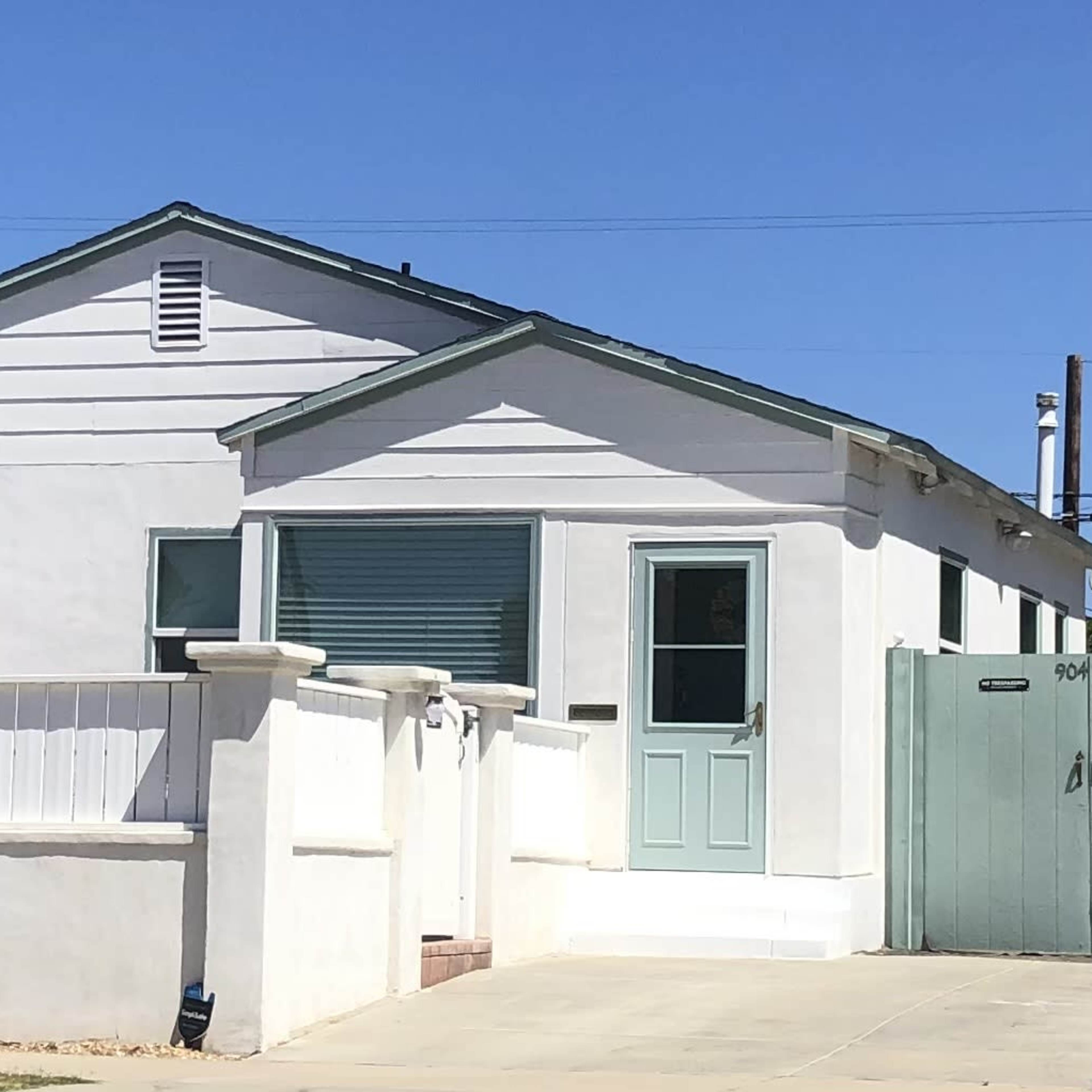 A white, single-story house with green trim and closed shutters, situated on a corner lot with a fence.