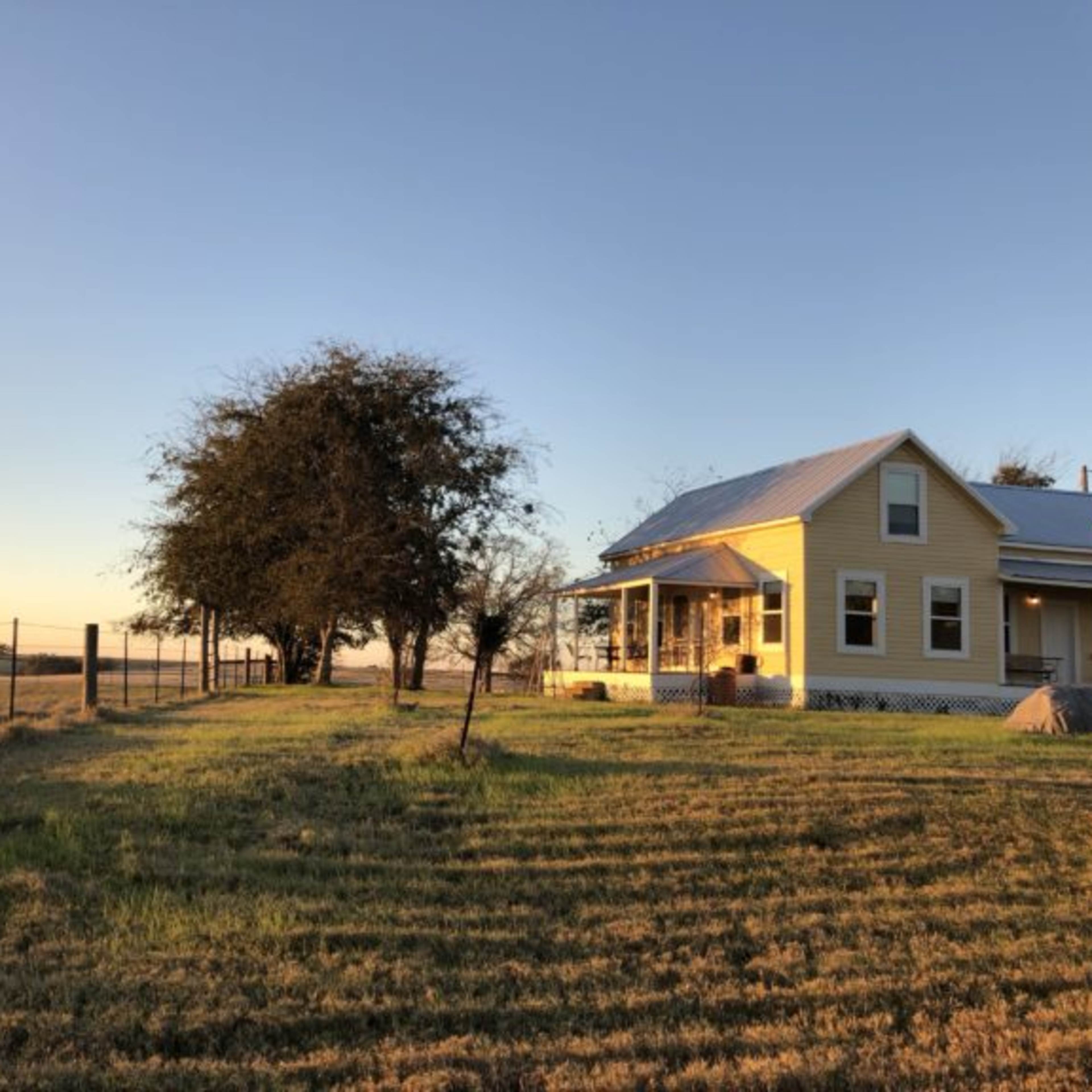 A yellow farmhouse is situated on a grassy hill, with a fence and a few trees in the background under a clear sky.