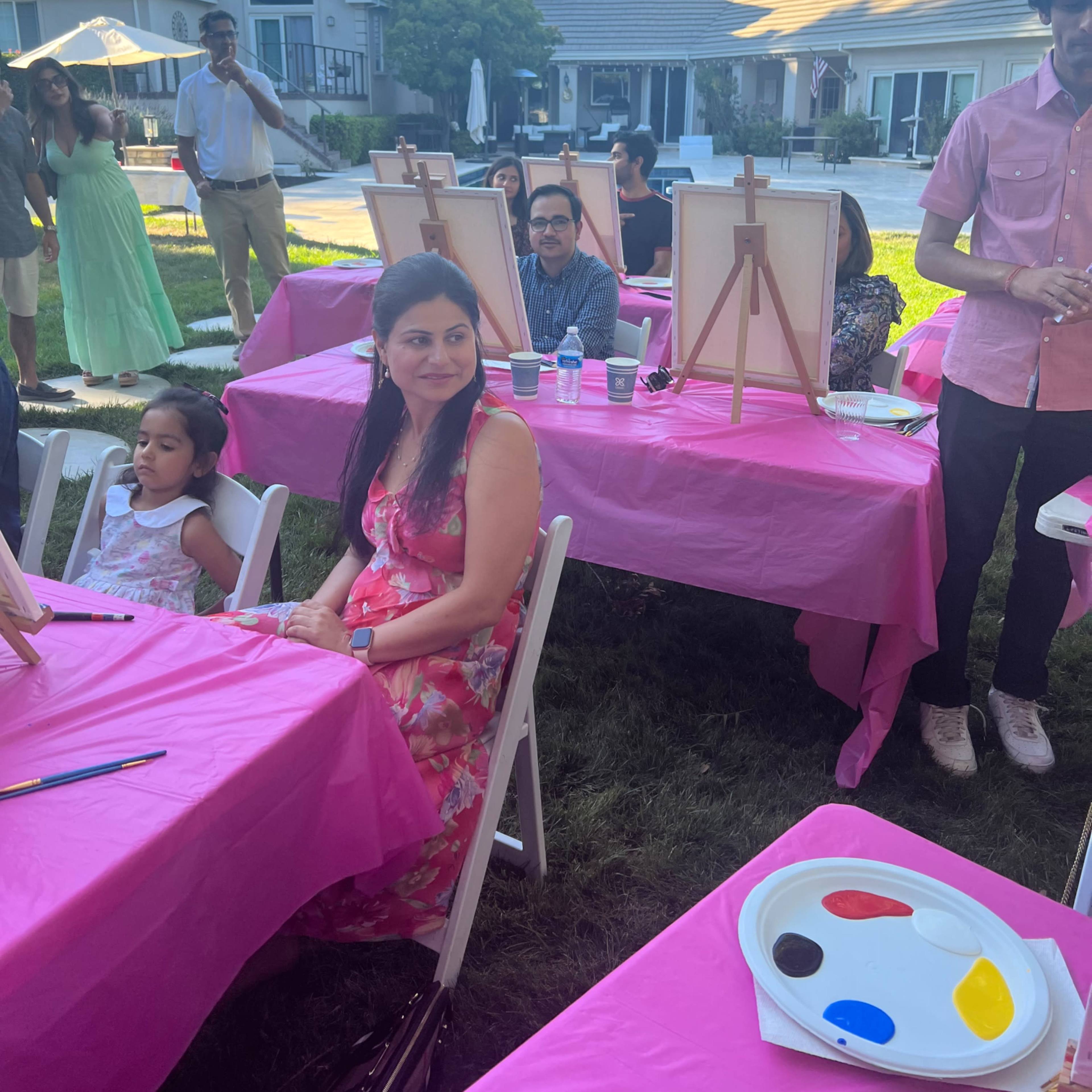 A group of people gathers around pink tables set up for a painting event in a backyard with a house in the background.