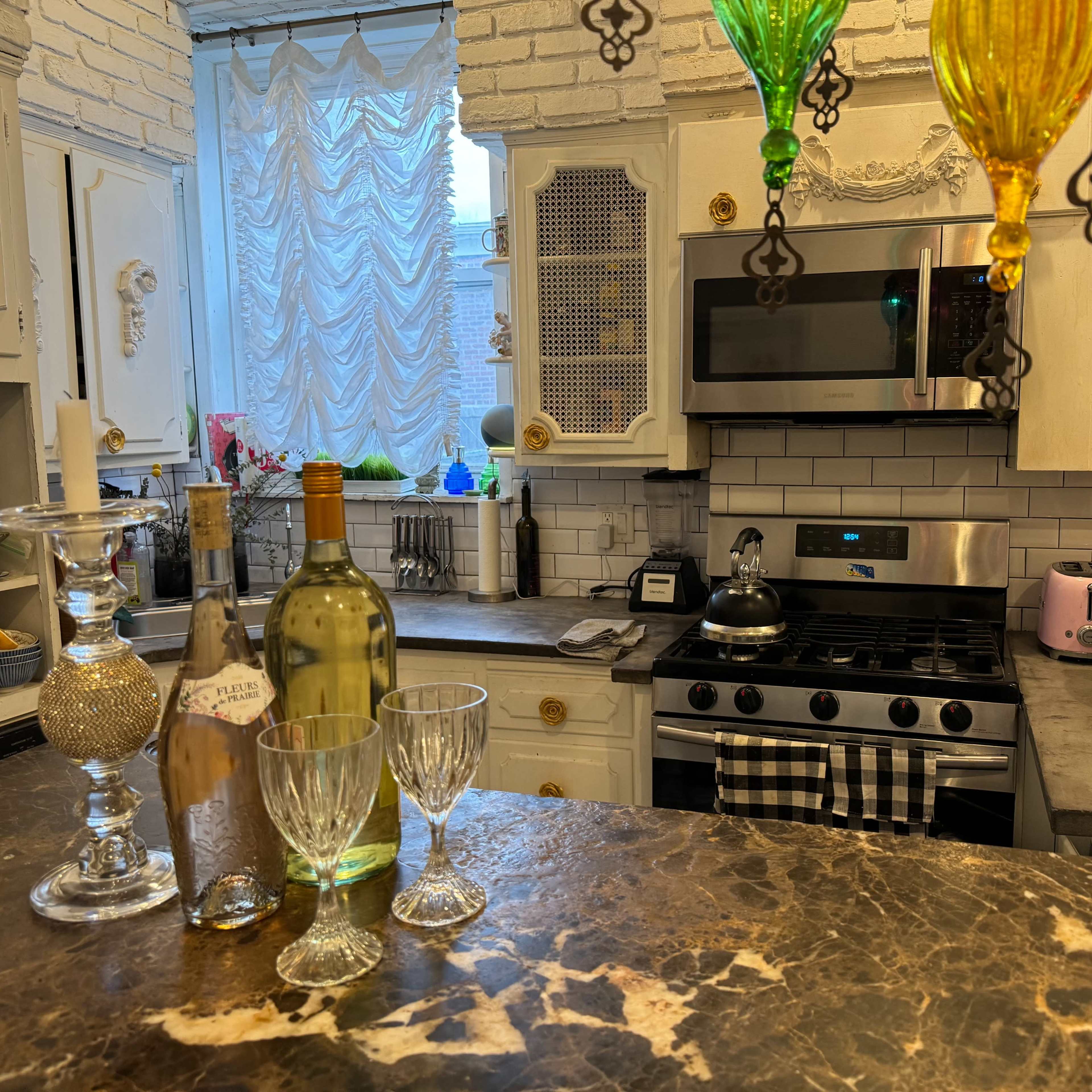 A cluttered kitchen features a marble countertop with bottles of wine, crystal glasses, and colorful pendant lights hanging above the stove and sink area.