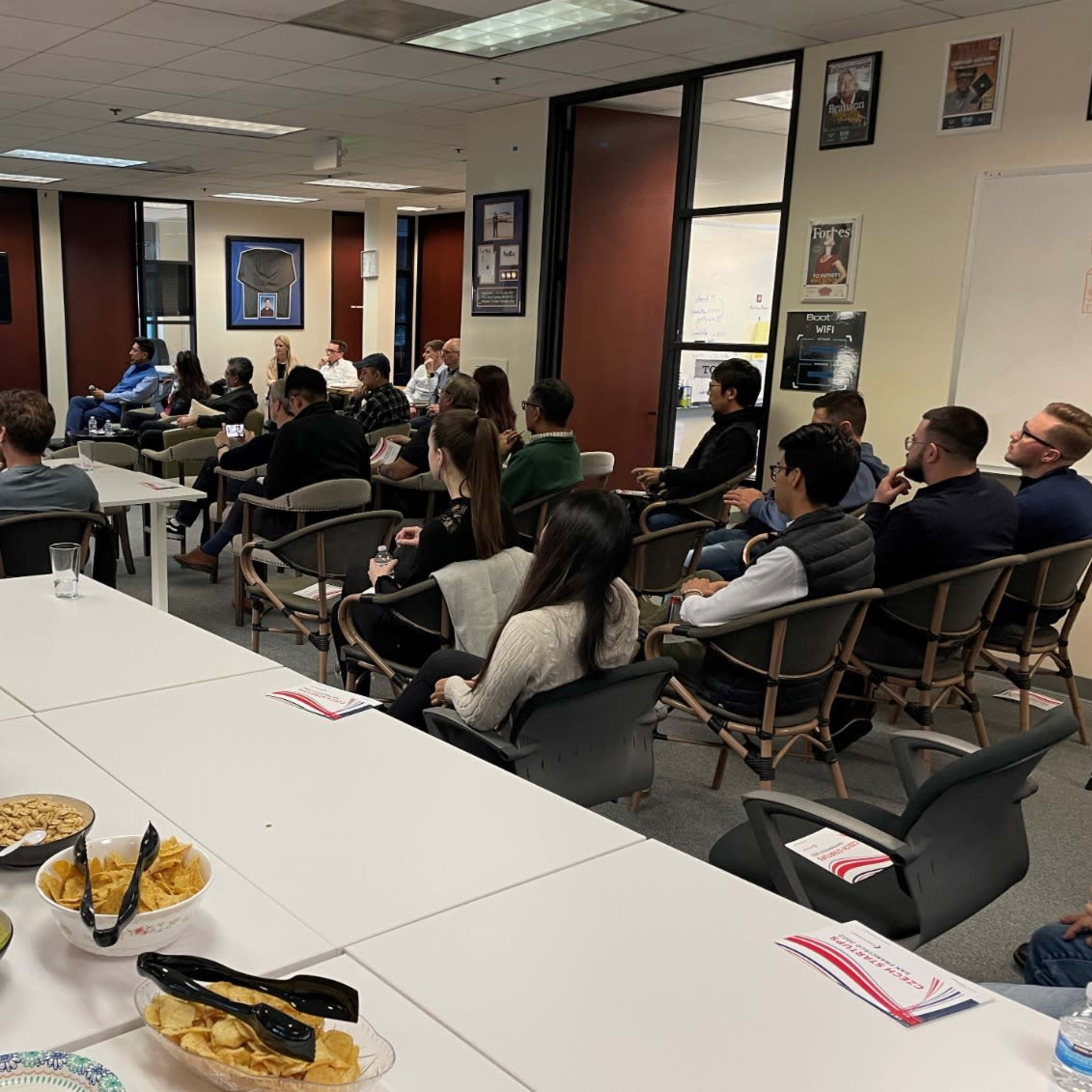 A group of people is seated in a meeting room, attentively watching a presentation while snacks and beverages are arranged on a table.