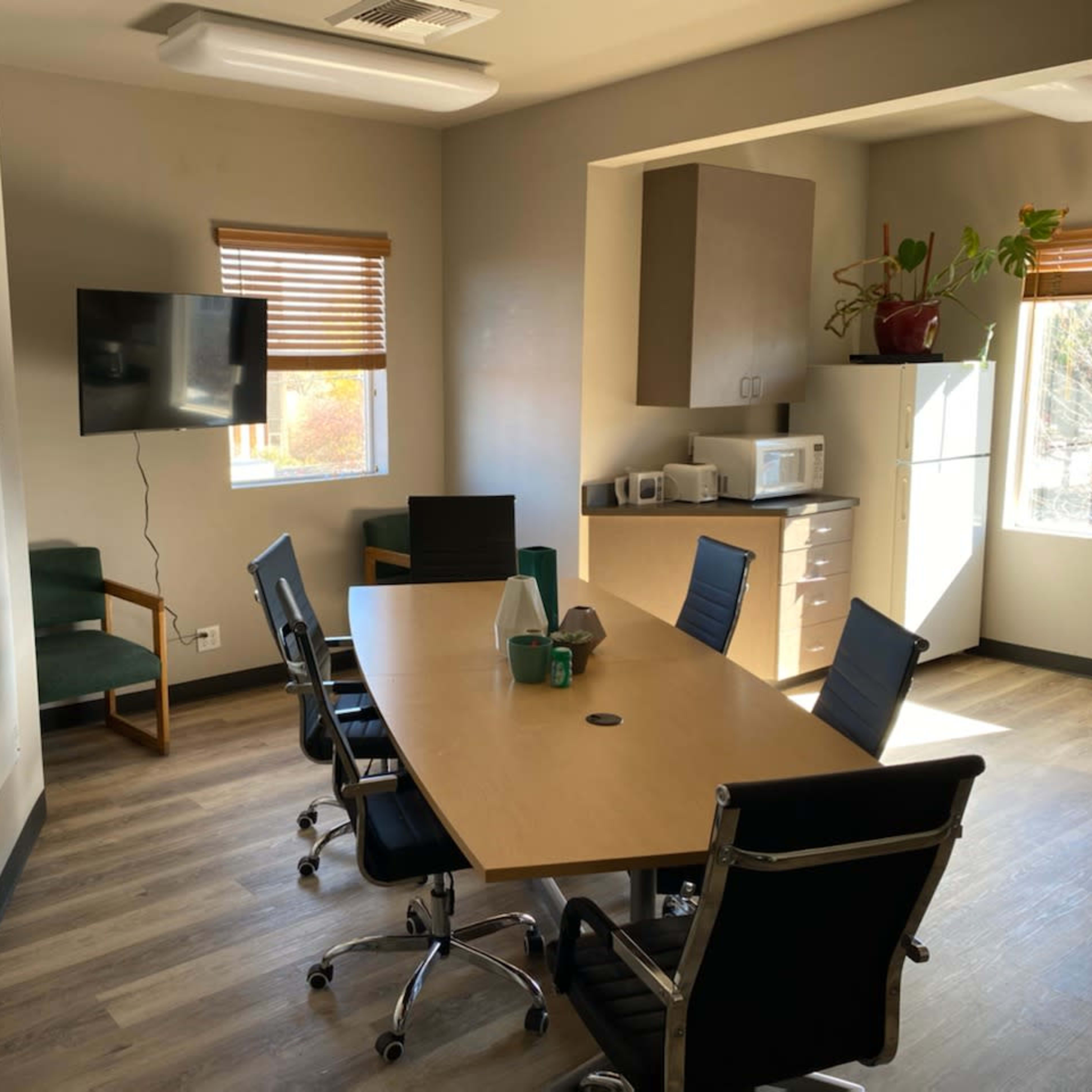 A conference room with a large table surrounded by black chairs, a television mounted on the wall, and a kitchenette in the background.