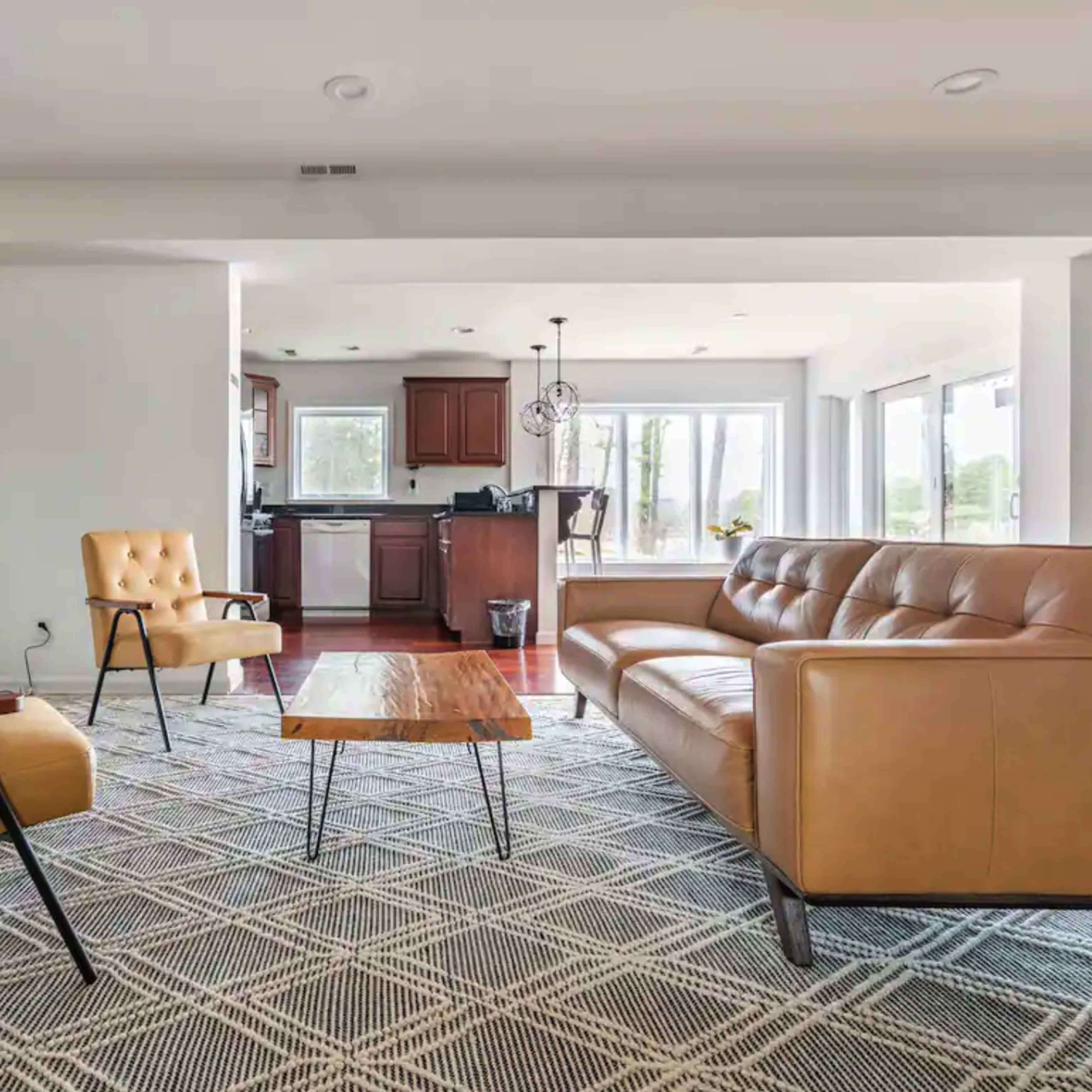 A modern living room with a tan leather couch, a wooden coffee table, and two armchairs, connected to a kitchen area with dark wood cabinets and large windows.