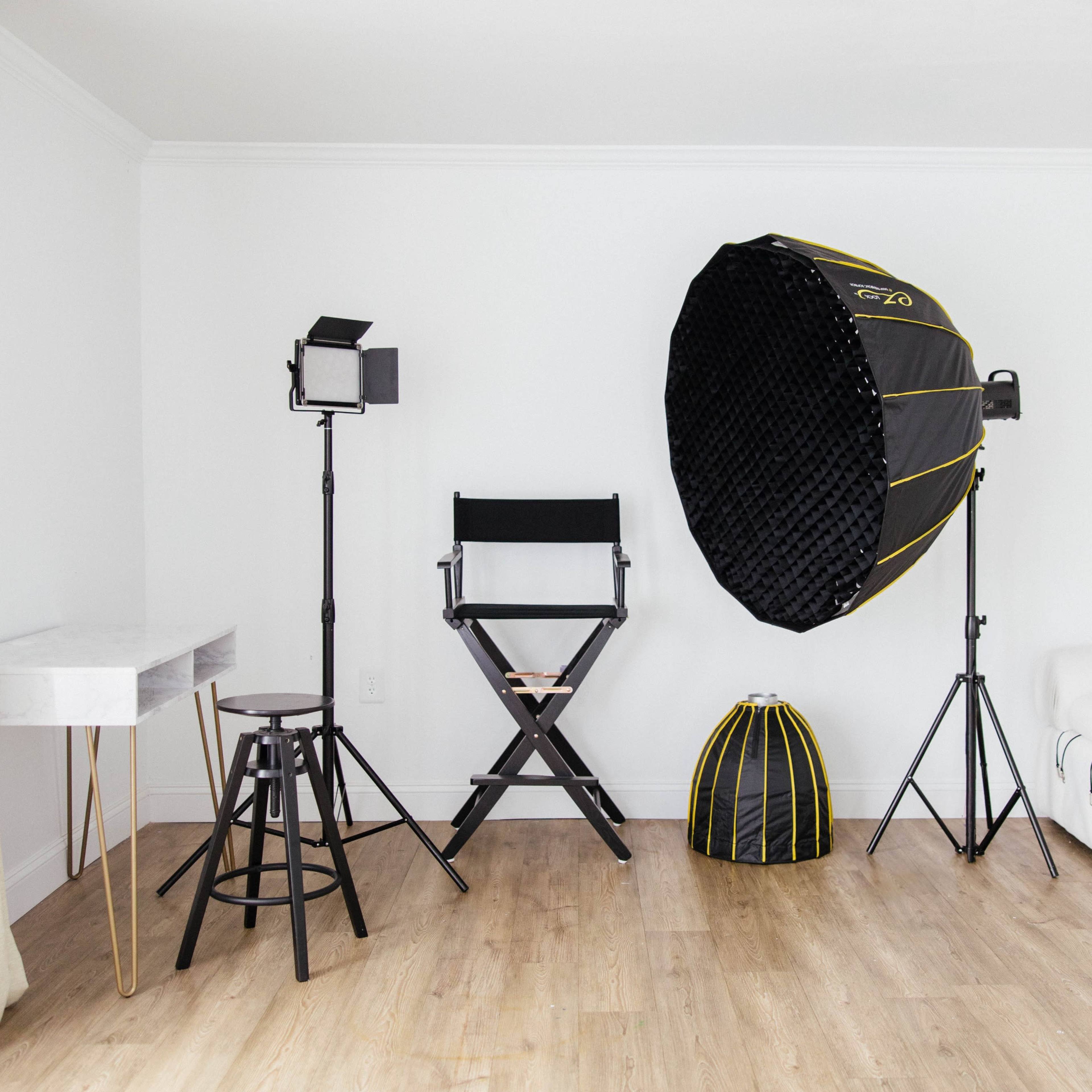 The scene shows a minimalist studio setting with a director's chair, lighting equipment, a large softbox, and a white sofa against a light-colored wall.
