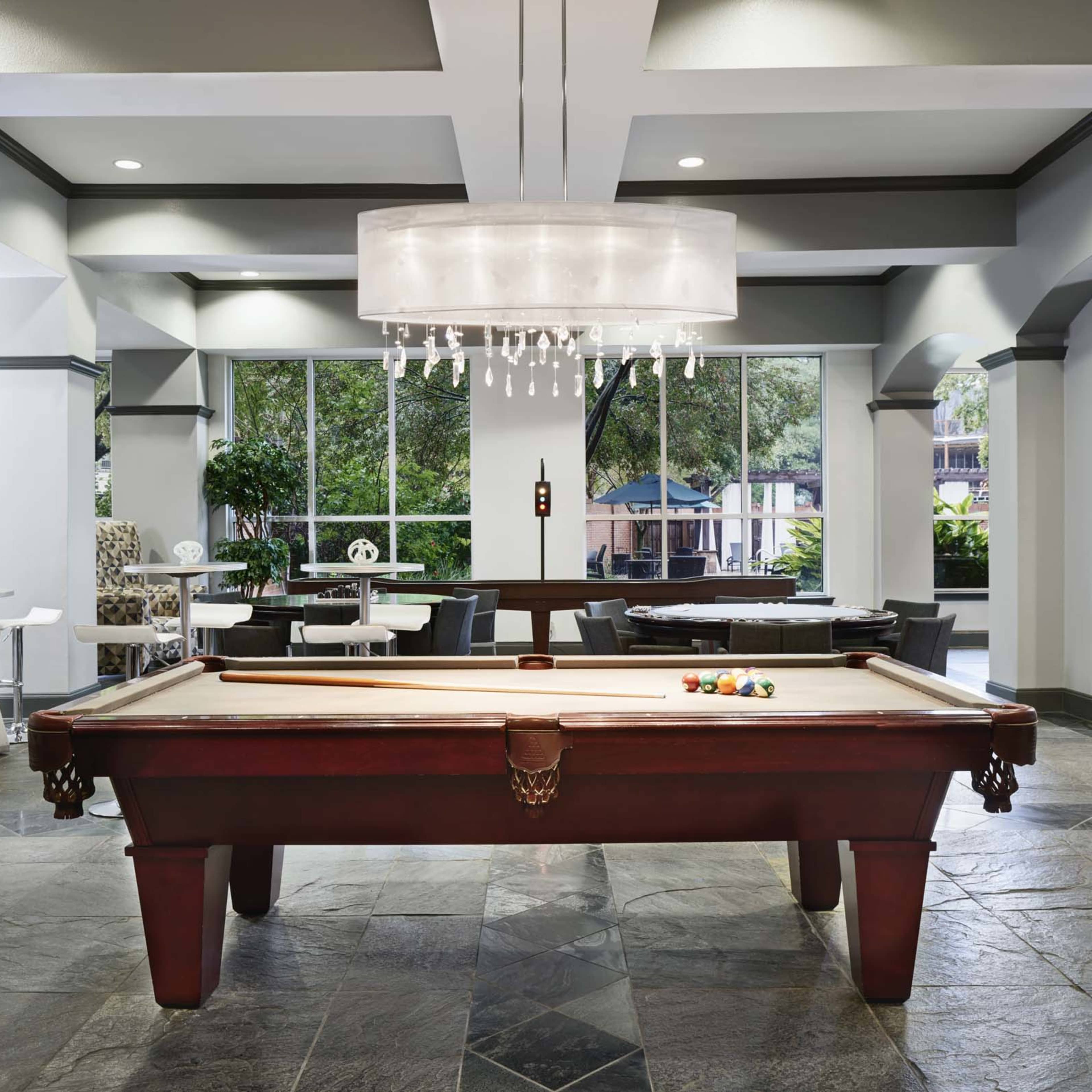 A modern lounge area featuring a wooden pool table at the center, surrounded by various seating arrangements and large windows.