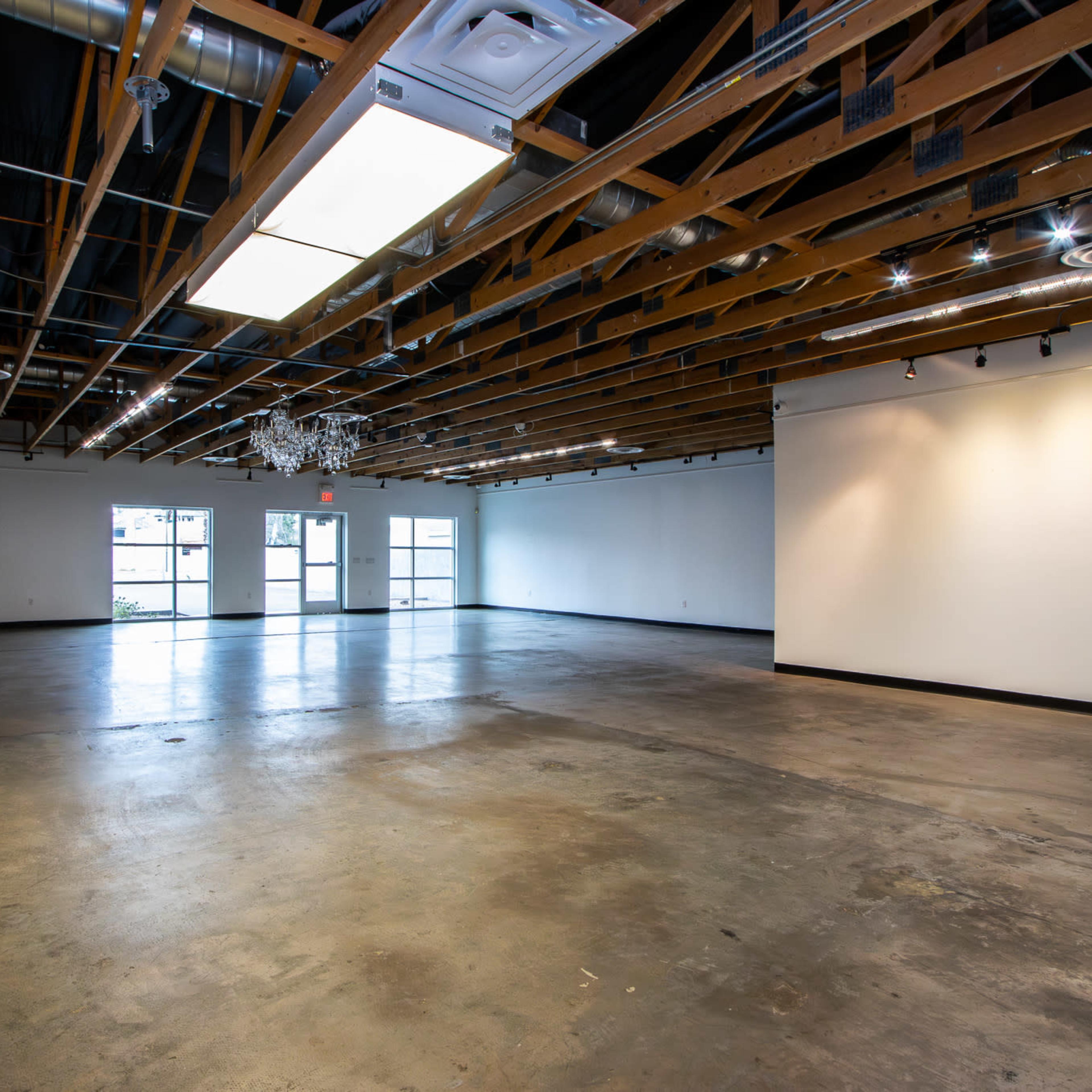 An empty loft-like space with exposed wooden beams, concrete flooring, and large windows at one end.