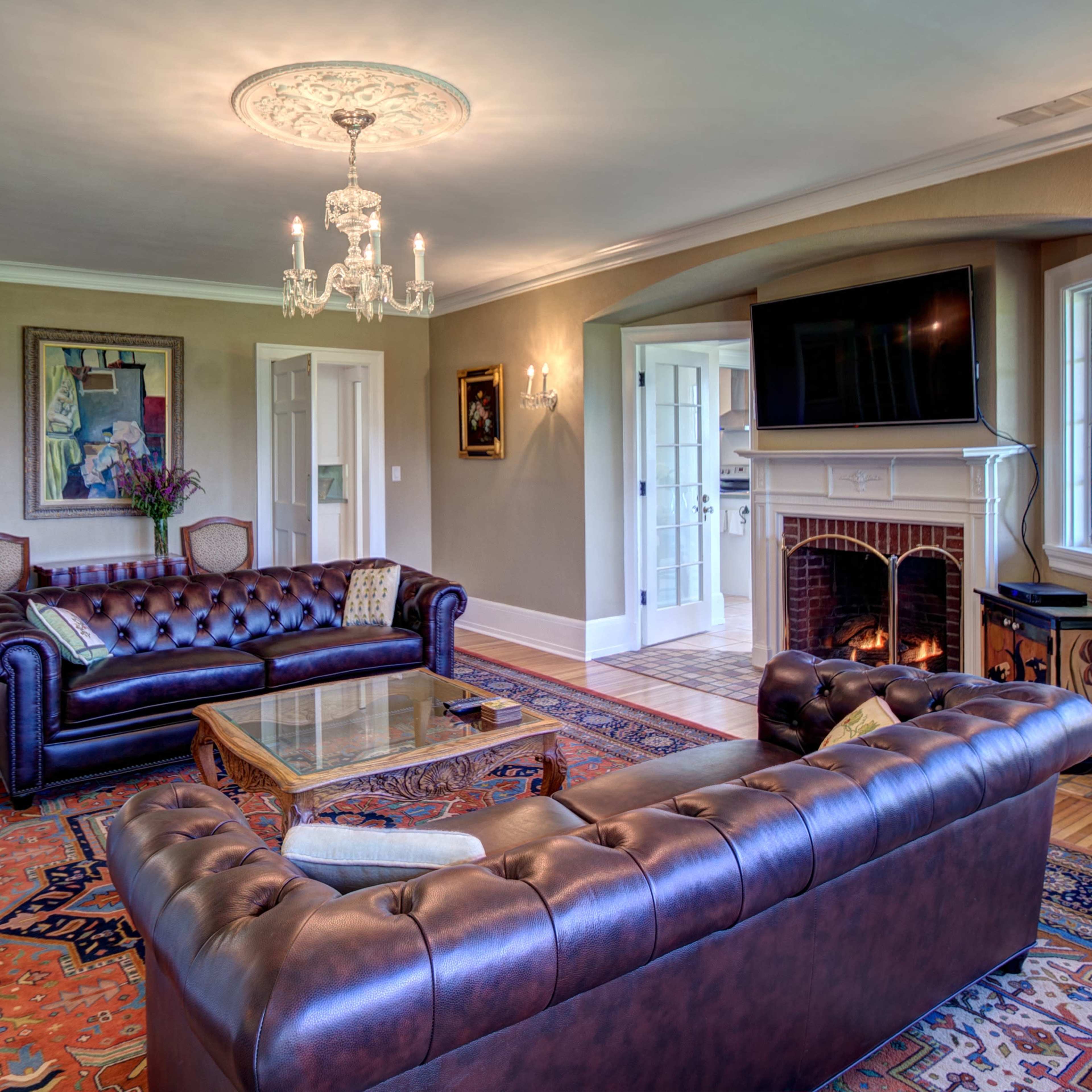 A spacious living room with a leather sofa, armchairs, a coffee table, a fireplace, and large windows overlooking a garden.