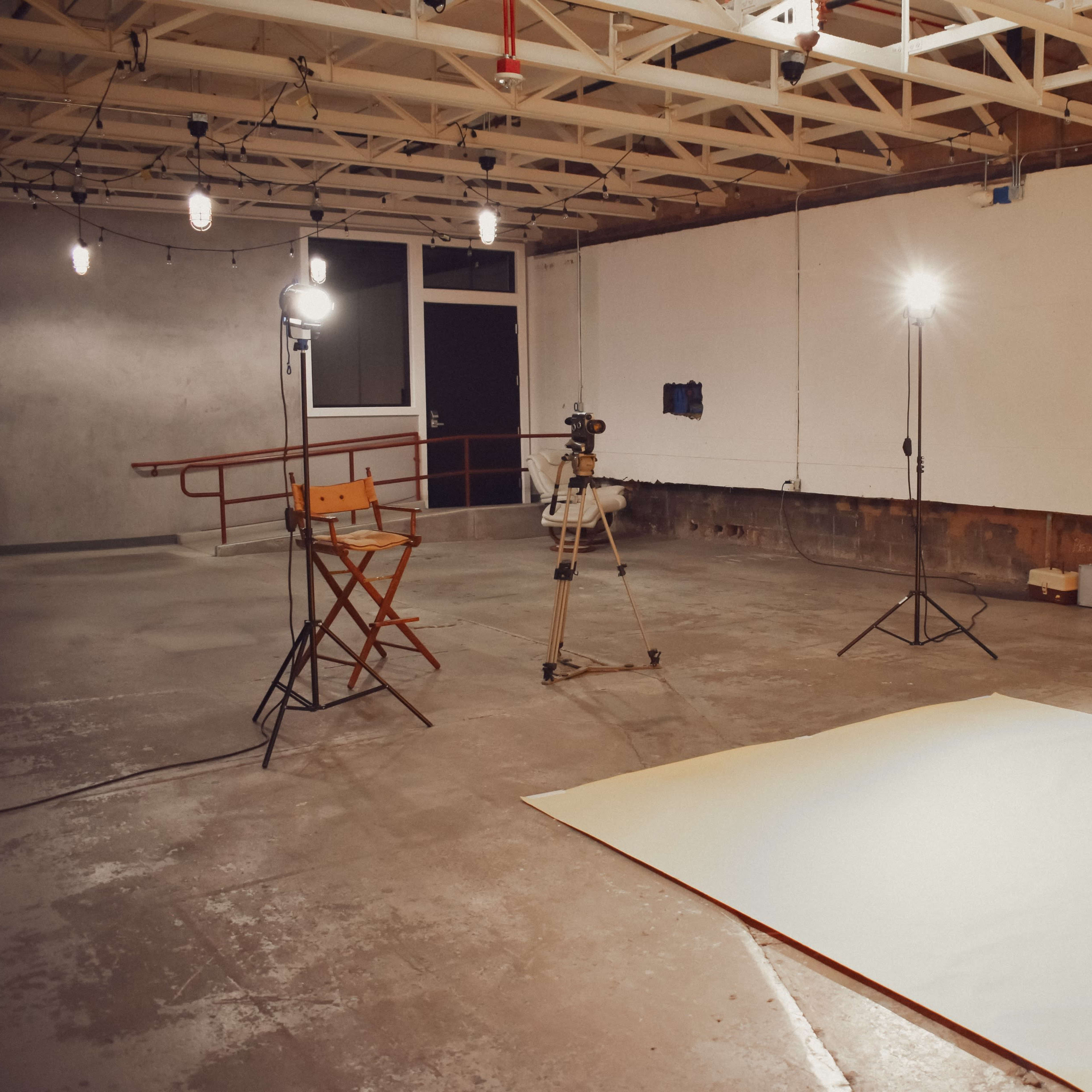 A photography studio is set up with a large orange backdrop, a director's chair, and several lighting equipment positioned around a concrete floor.