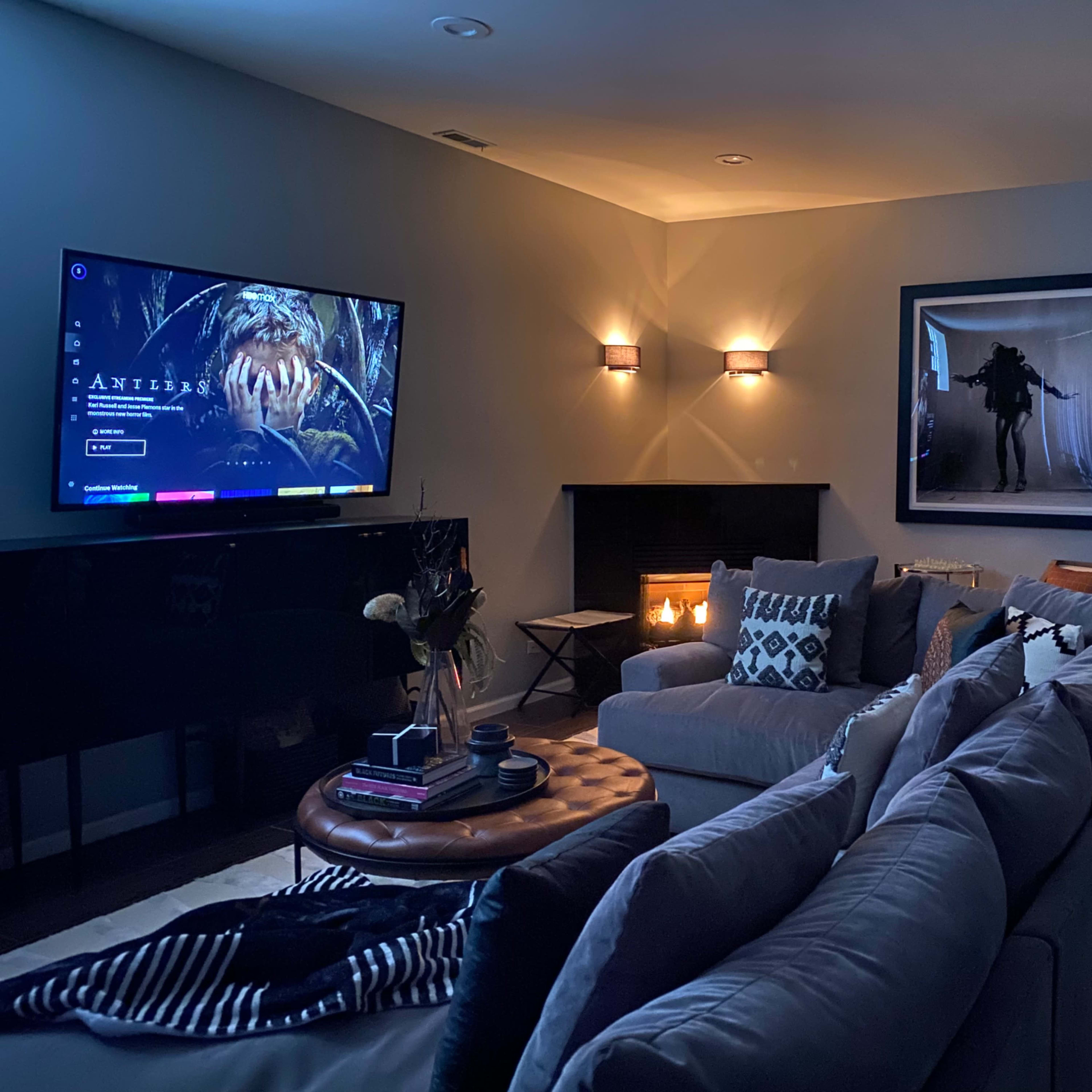 A cozy living room features a large television displaying a digital interface, a sectional sofa, a round coffee table, and wall-mounted lights illuminating the space.