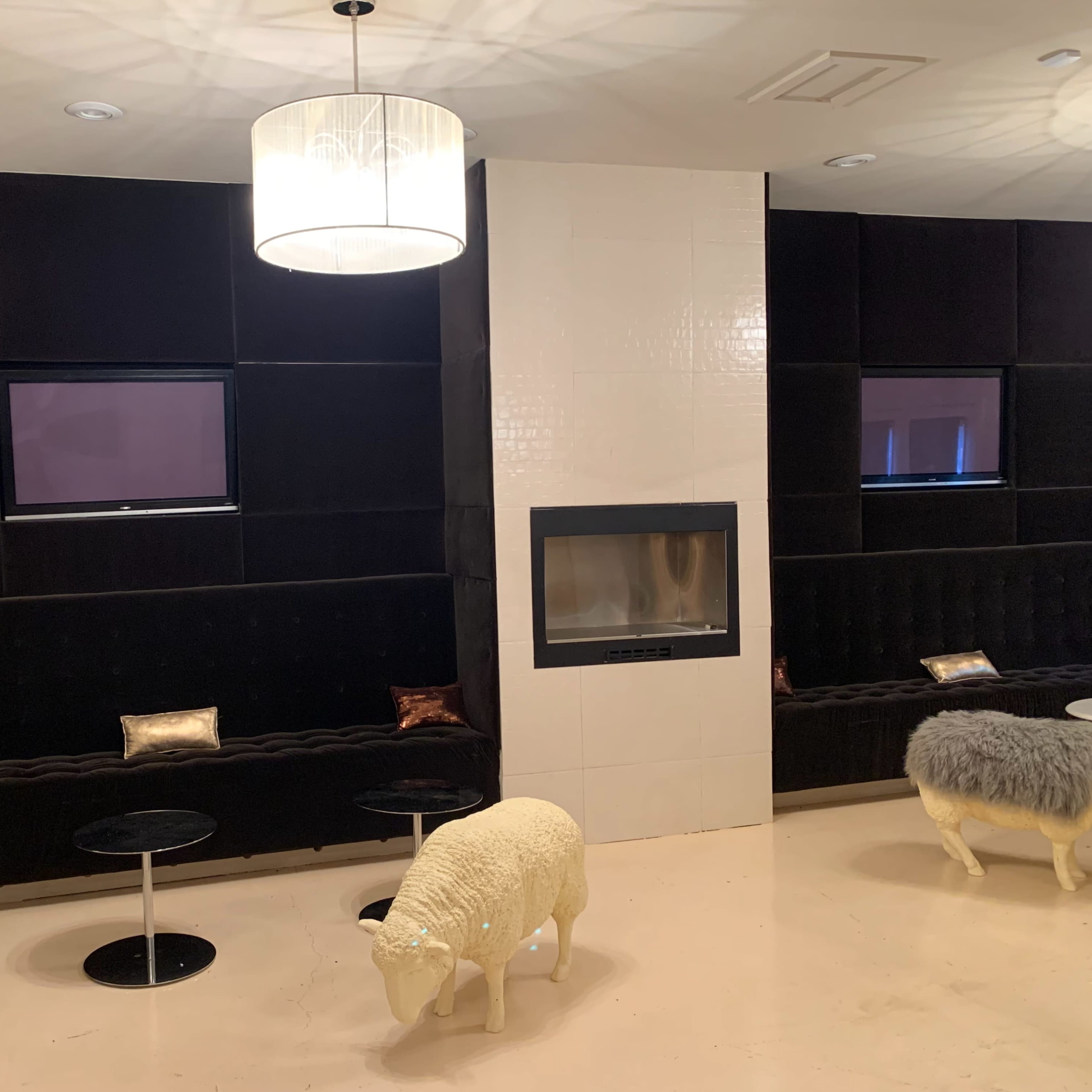 A modern lounge area with black tufted seating, a white fireplace, a large light fixture, and two small tables, accompanied by decorative sheep sculptures.