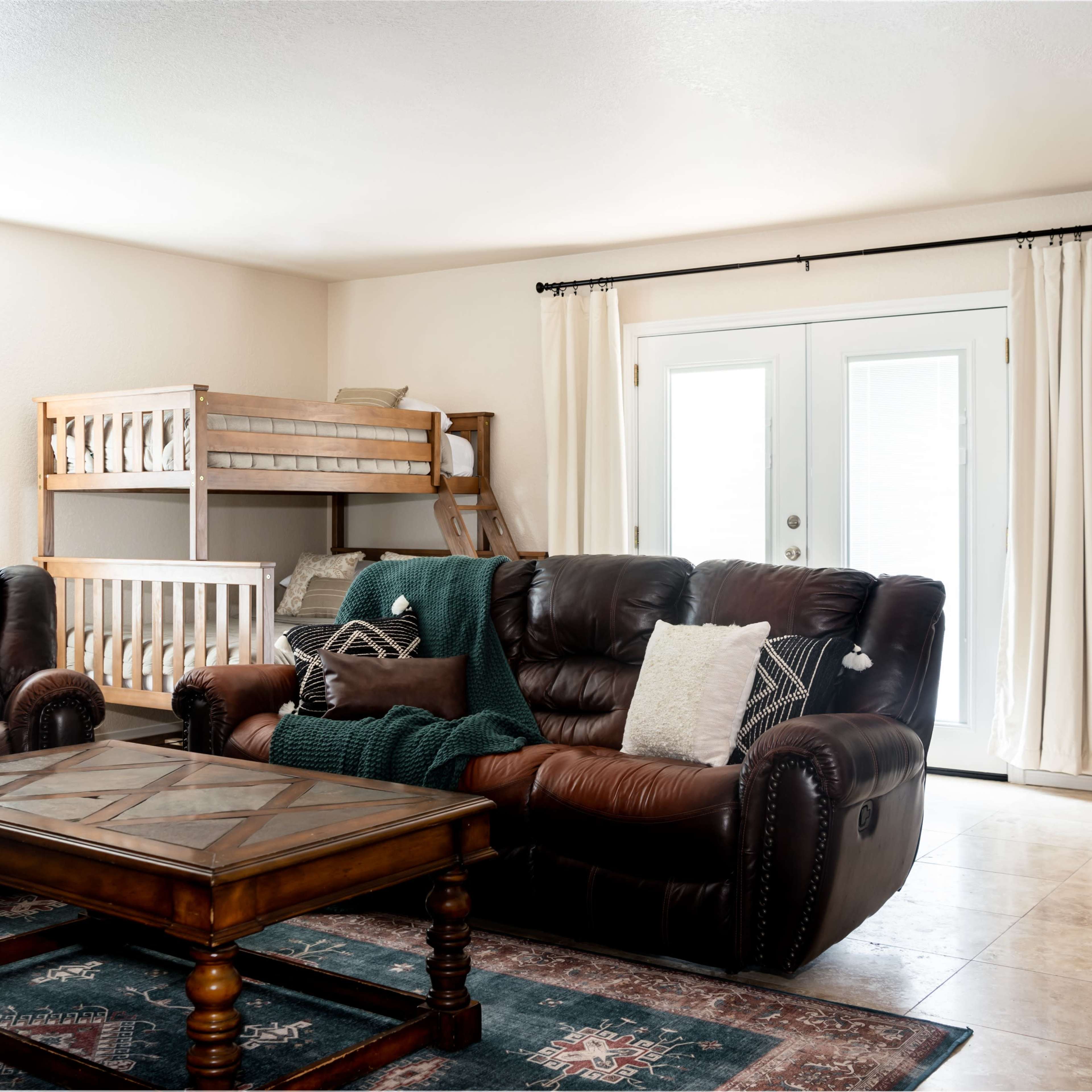 A cozy living room features two leather sofas facing a wooden coffee table, with a bunk bed in the background and sliding glass doors leading outside.