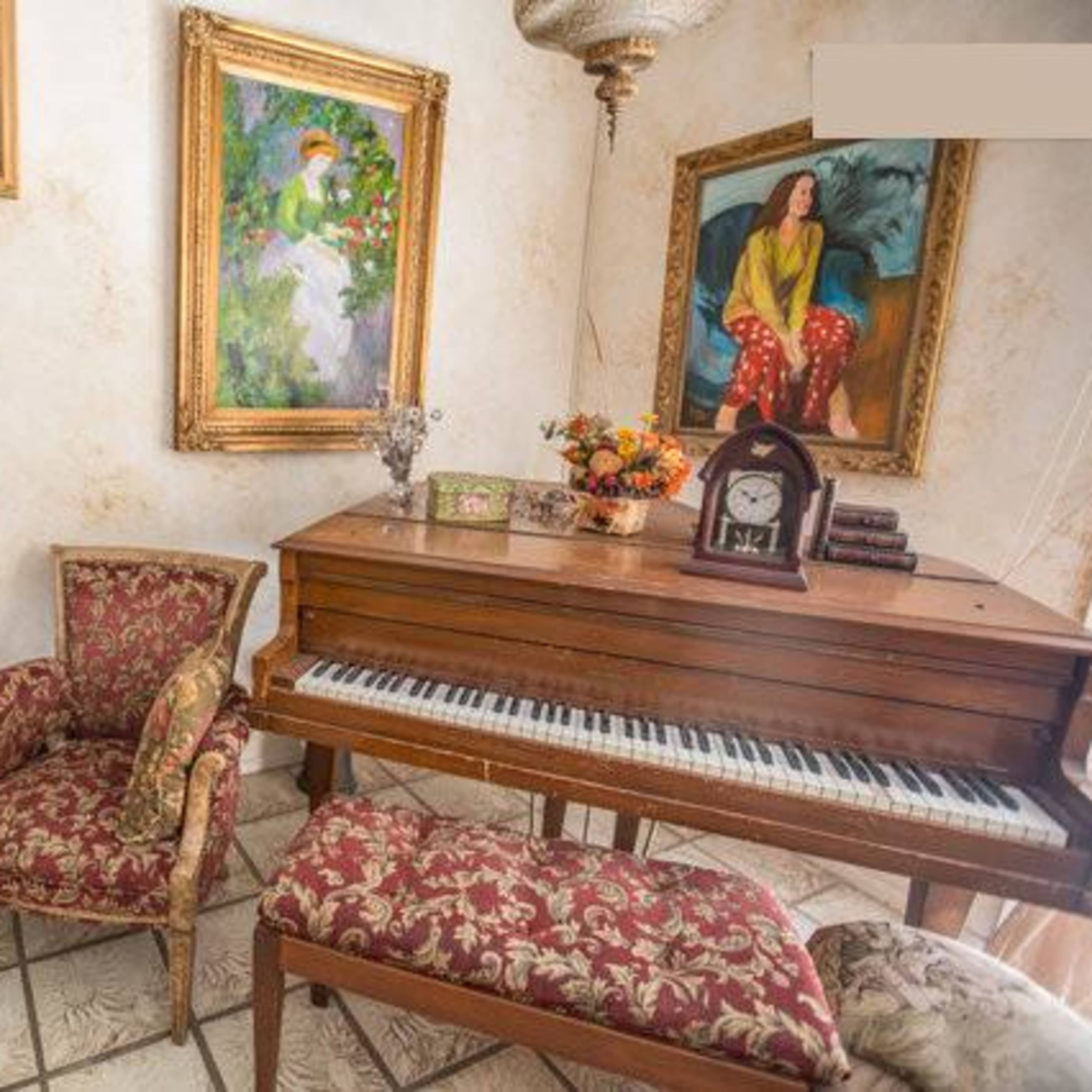A cozy room featuring a wooden piano, patterned chairs, a small round table, and several framed paintings on the walls.