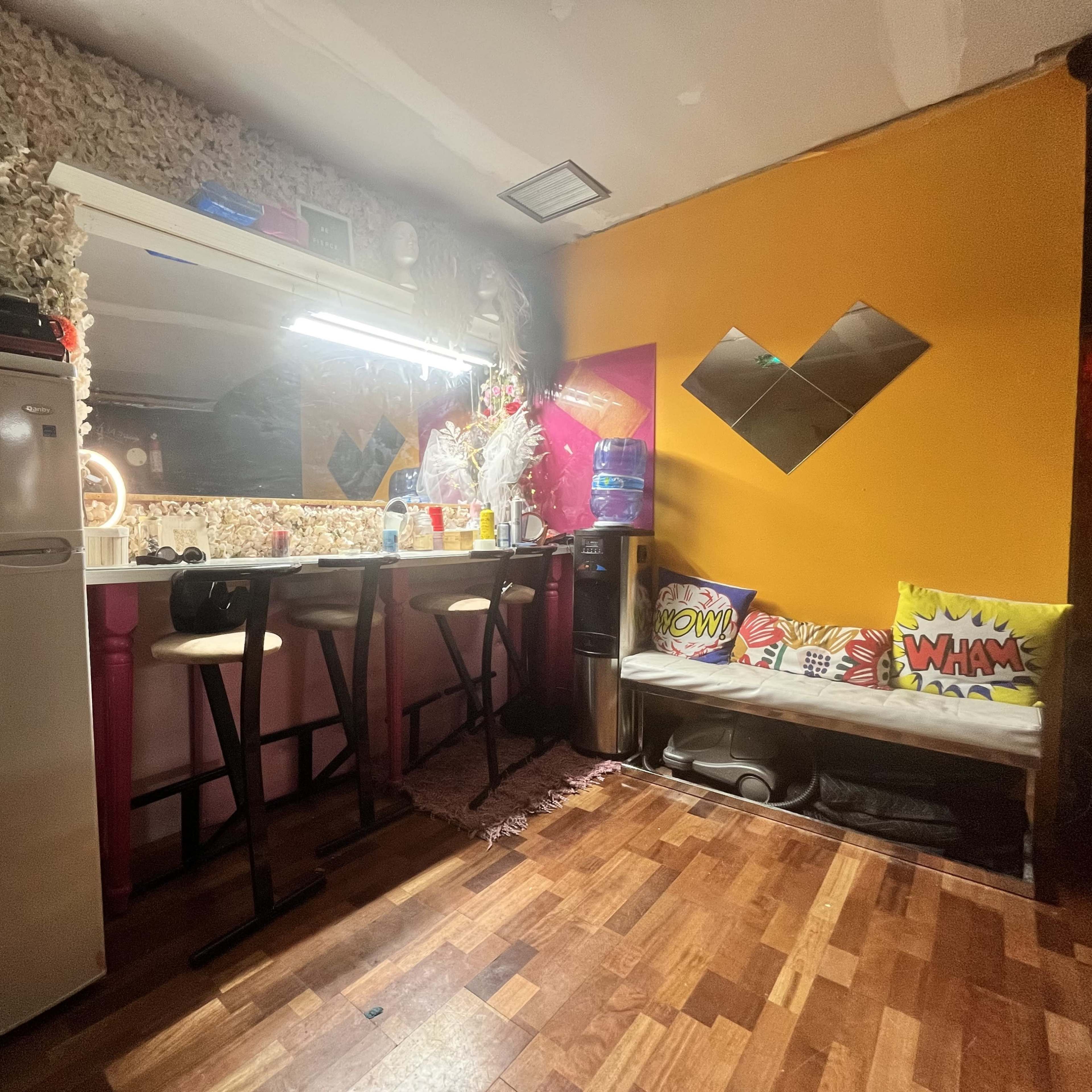 A small kitchen area with a bar counter, stools, refrigerator, and a colorful wall featuring decorative cushions.