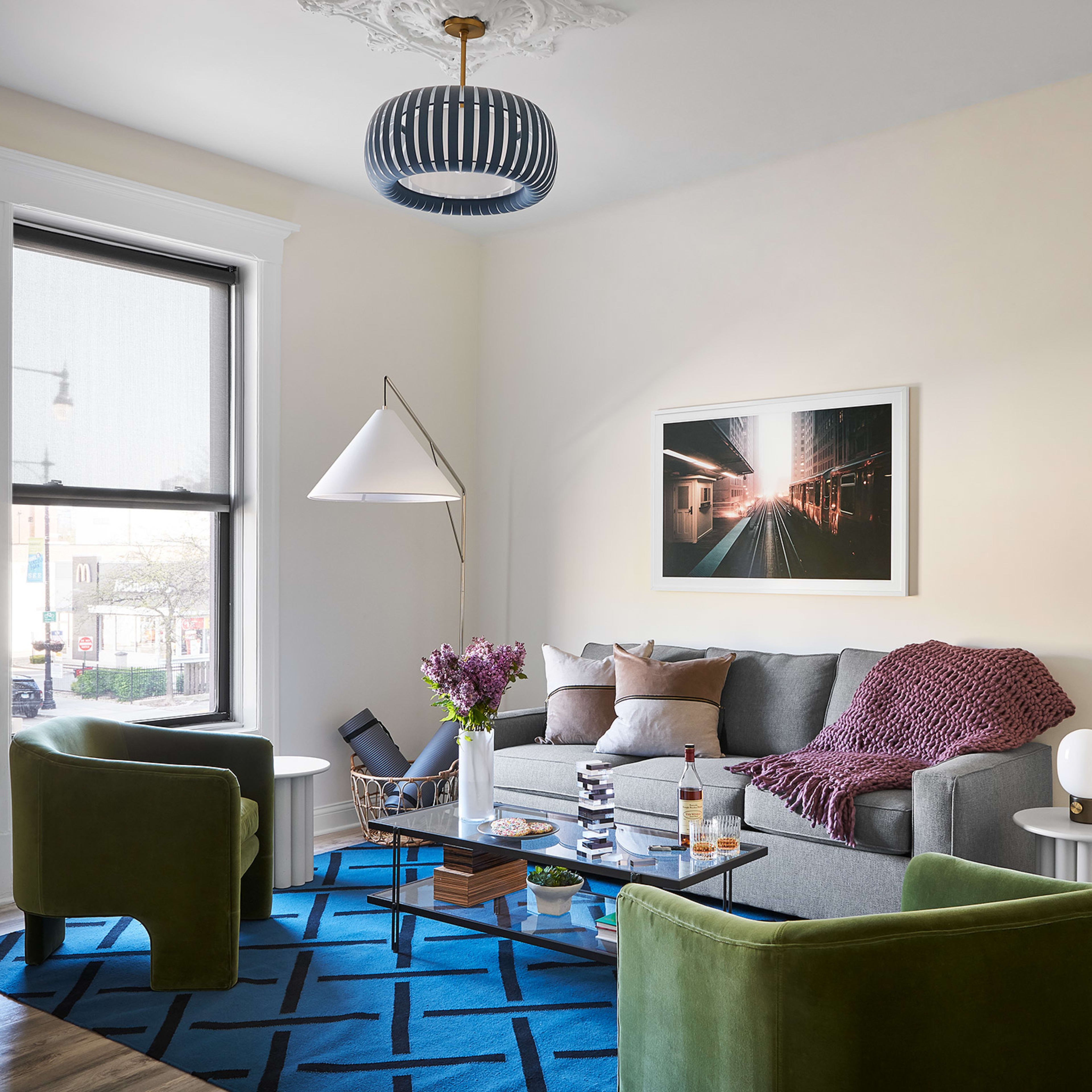 A modern living room features a gray sofa with patterned pillows, two green armchairs, a coffee table with decorative items, and a patterned rug under a ceiling light and artwork.