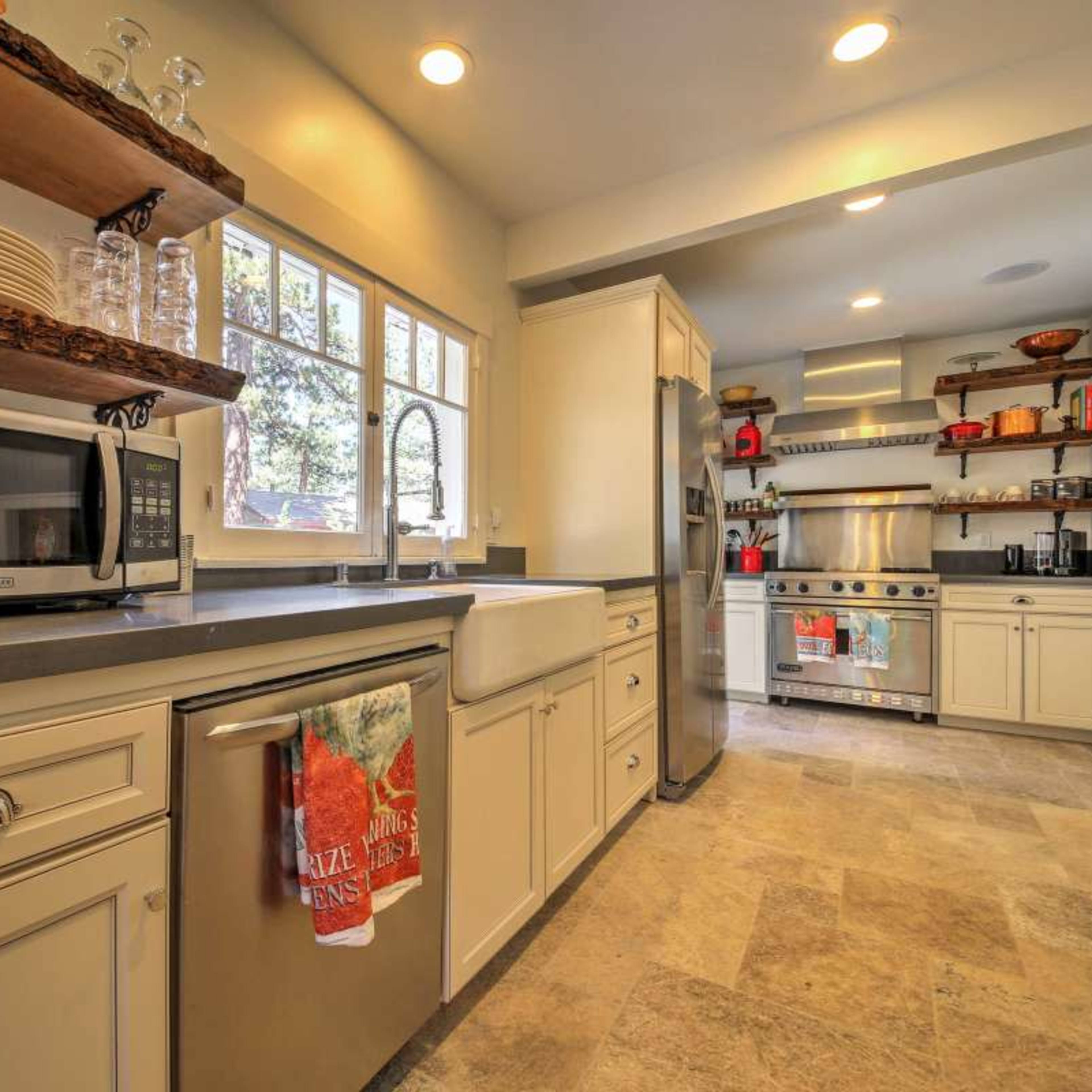 A modern kitchen featuring stainless steel appliances, a farmhouse sink, open shelving, and a combination of light cabinetry and stone flooring.