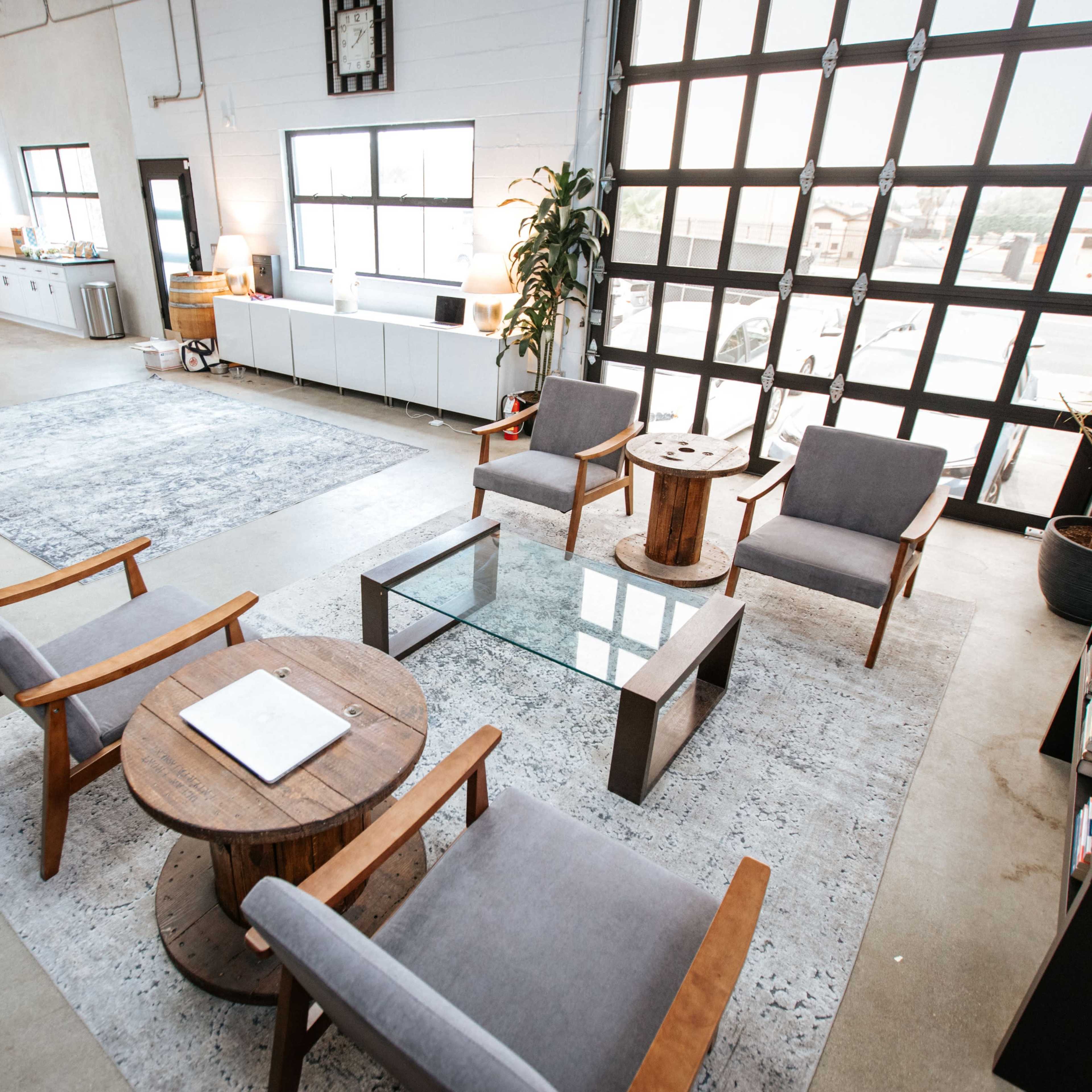 A spacious, modern living area with a kitchen in the background, featuring a seating arrangement of gray chairs and wooden tables on a light-colored rug.