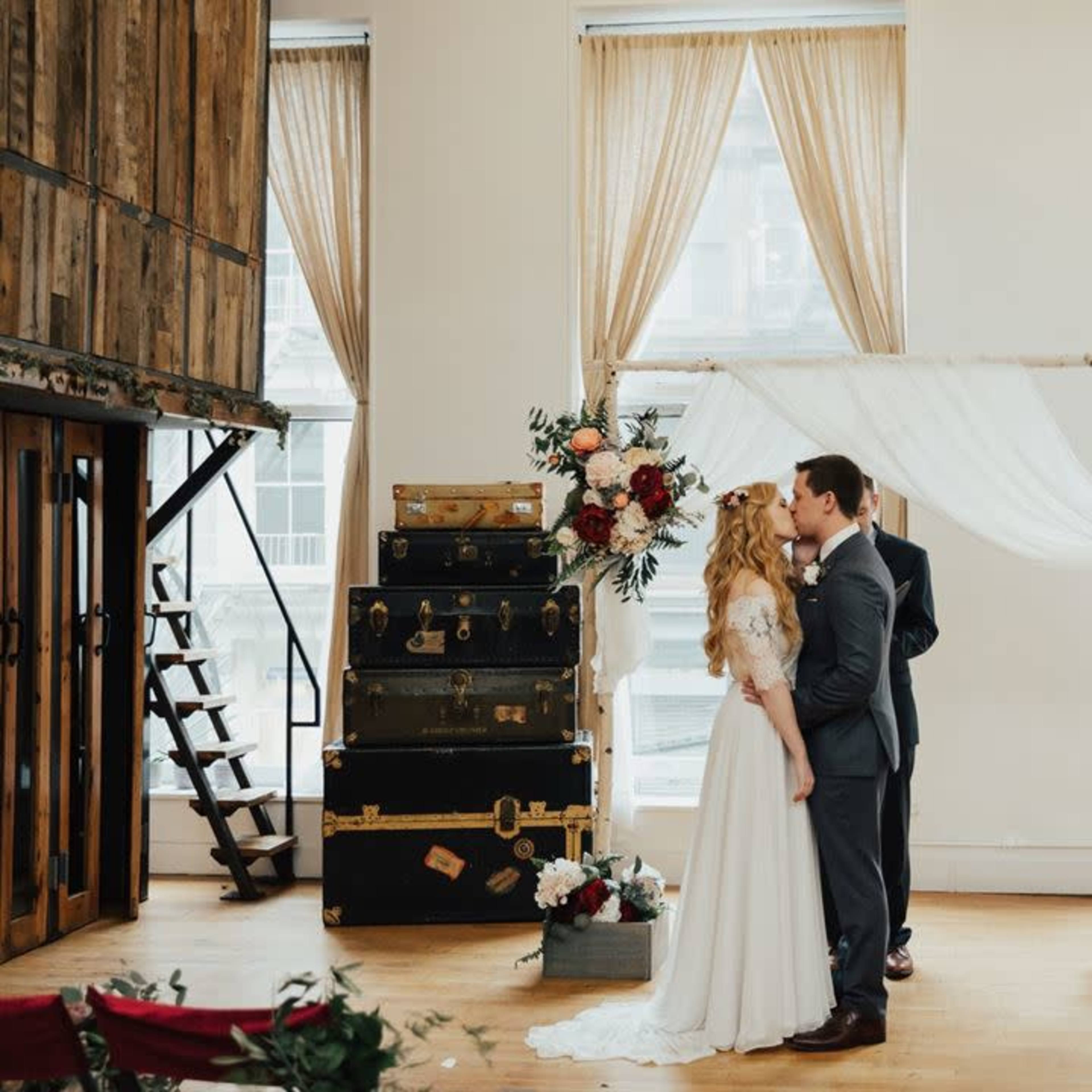 A bride and groom exchange vows under a floral arch in a decorated venue with large windows and vintage luggage in the background.