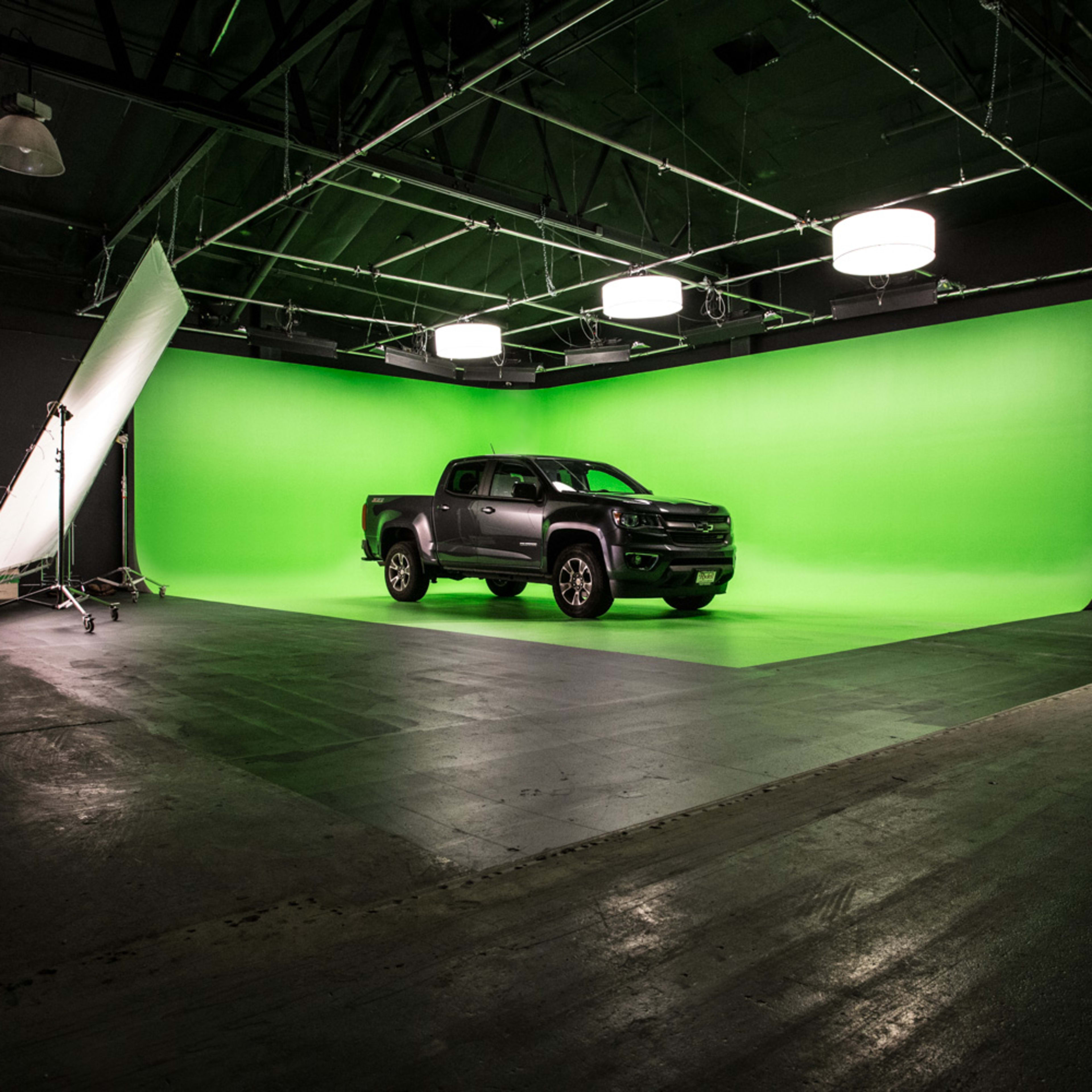 A black pickup truck is centered on a green screen backdrop in a spacious studio with industrial lighting.