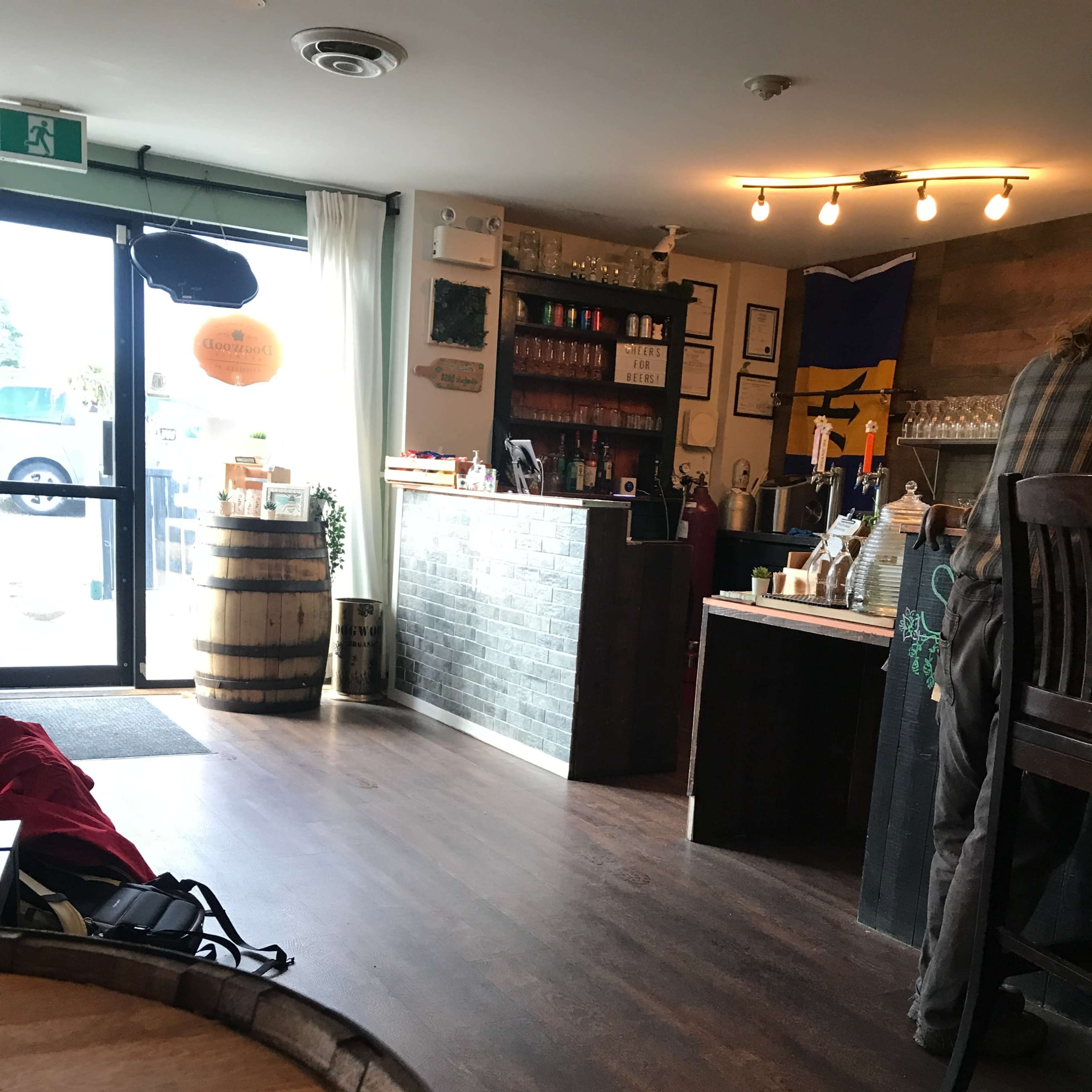 The interior of a bar or café with wooden flooring, a counter lined with bottles, and large windows letting in natural light.