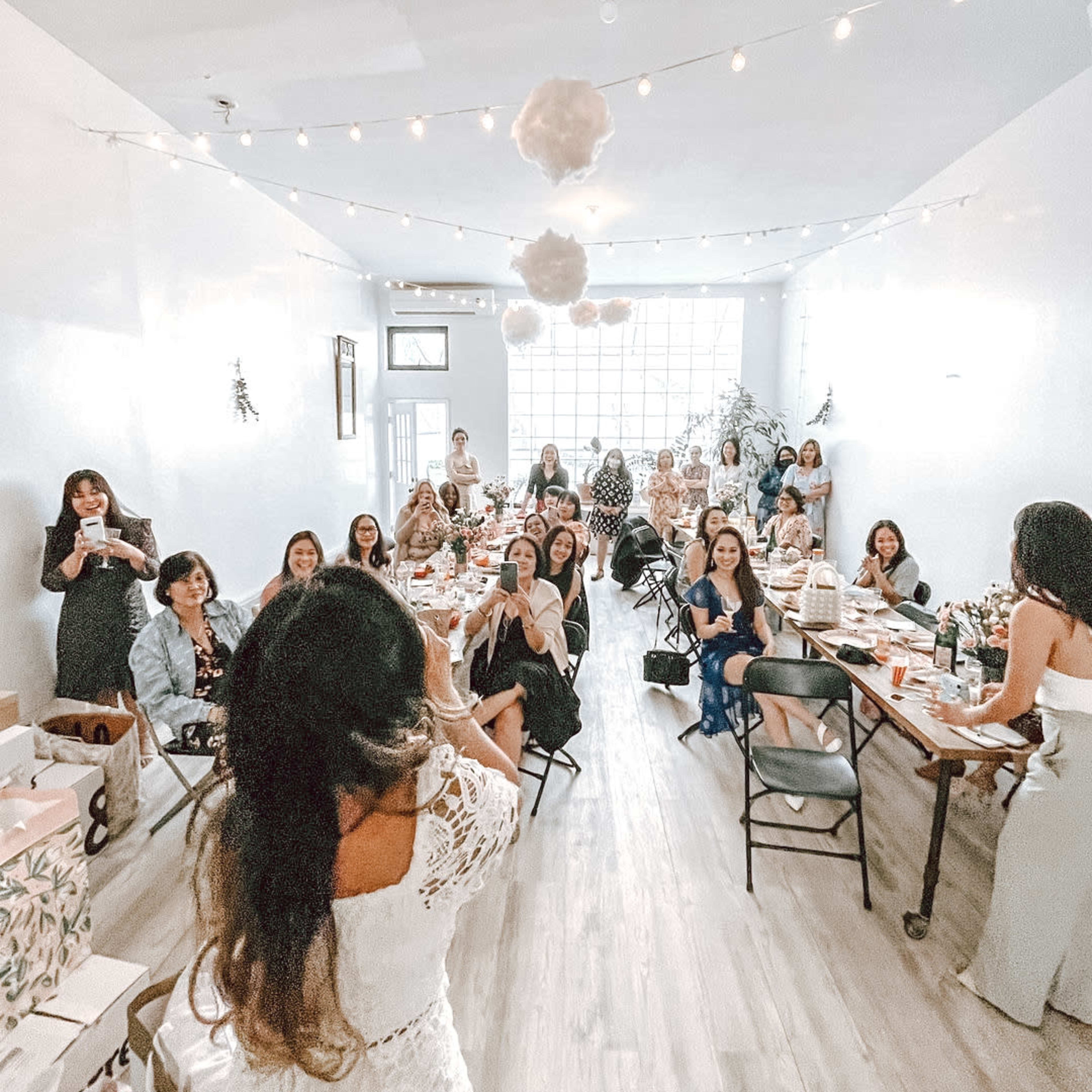 A group of women gathers in a brightly lit room decorated for a celebration, seated at tables with flowers and drinks.