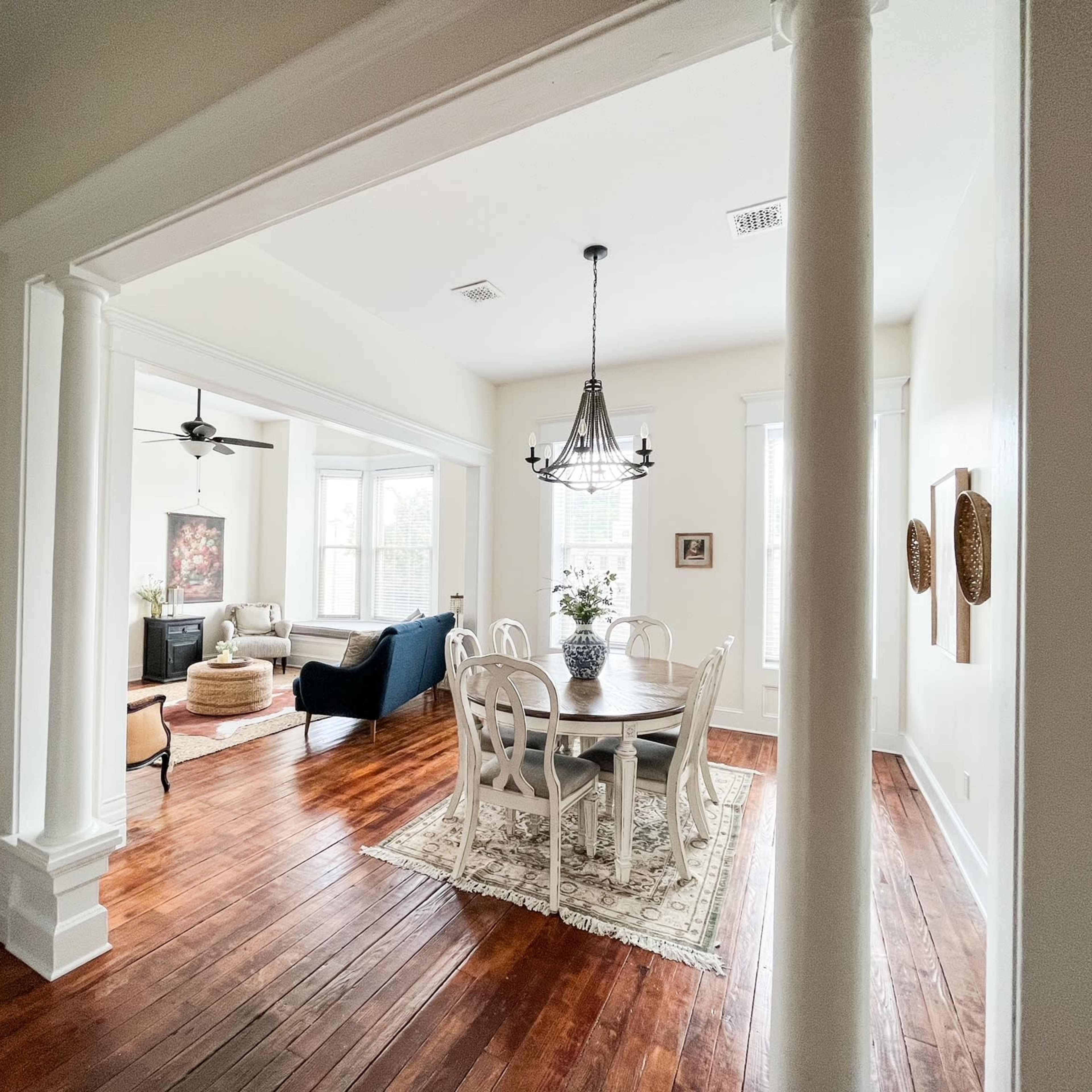 A room with a dining area featuring a round table and chairs, leading into a living space with a couch and large windows.