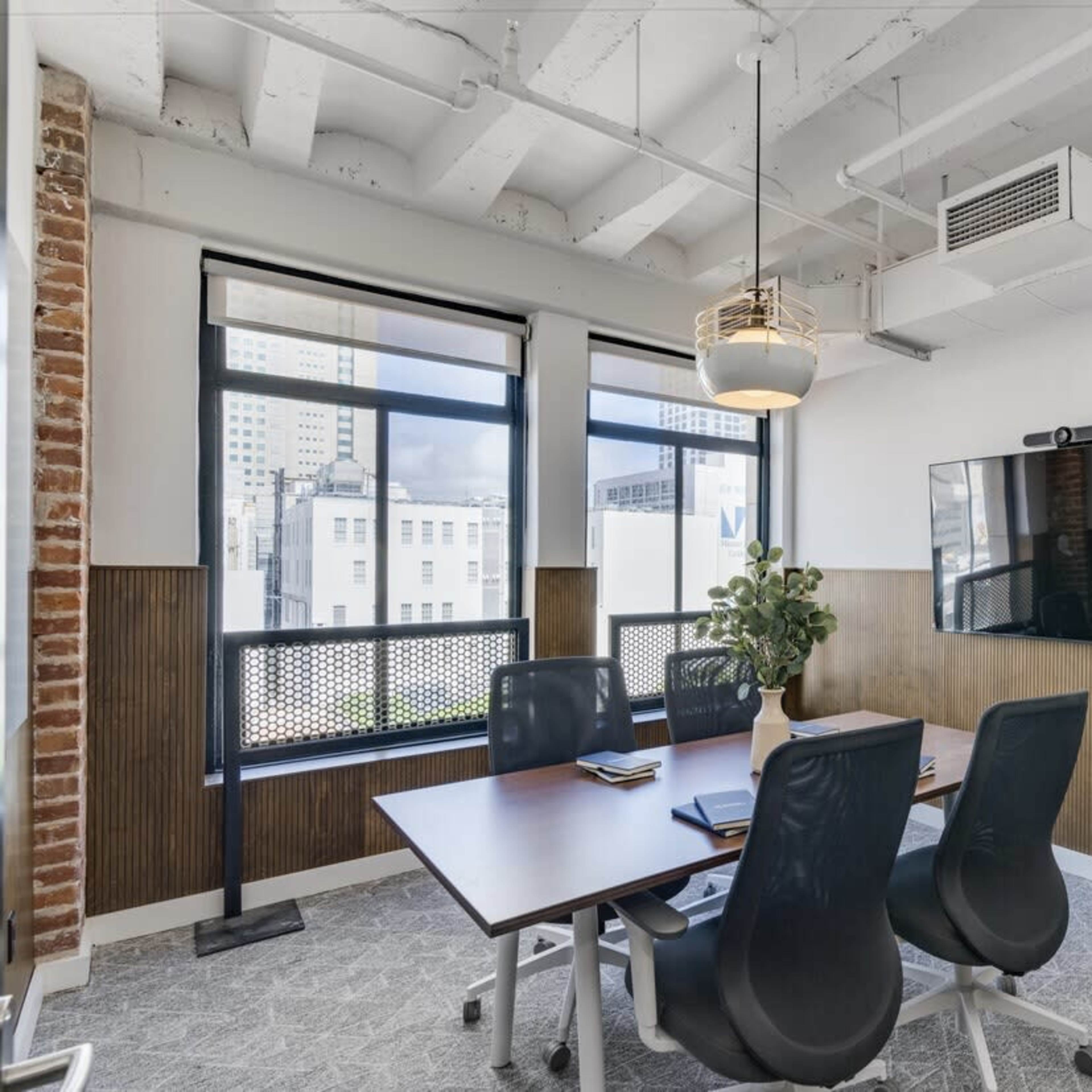 A modern conference room features a wooden table surrounded by chairs, a wall-mounted screen, and large windows with city views.