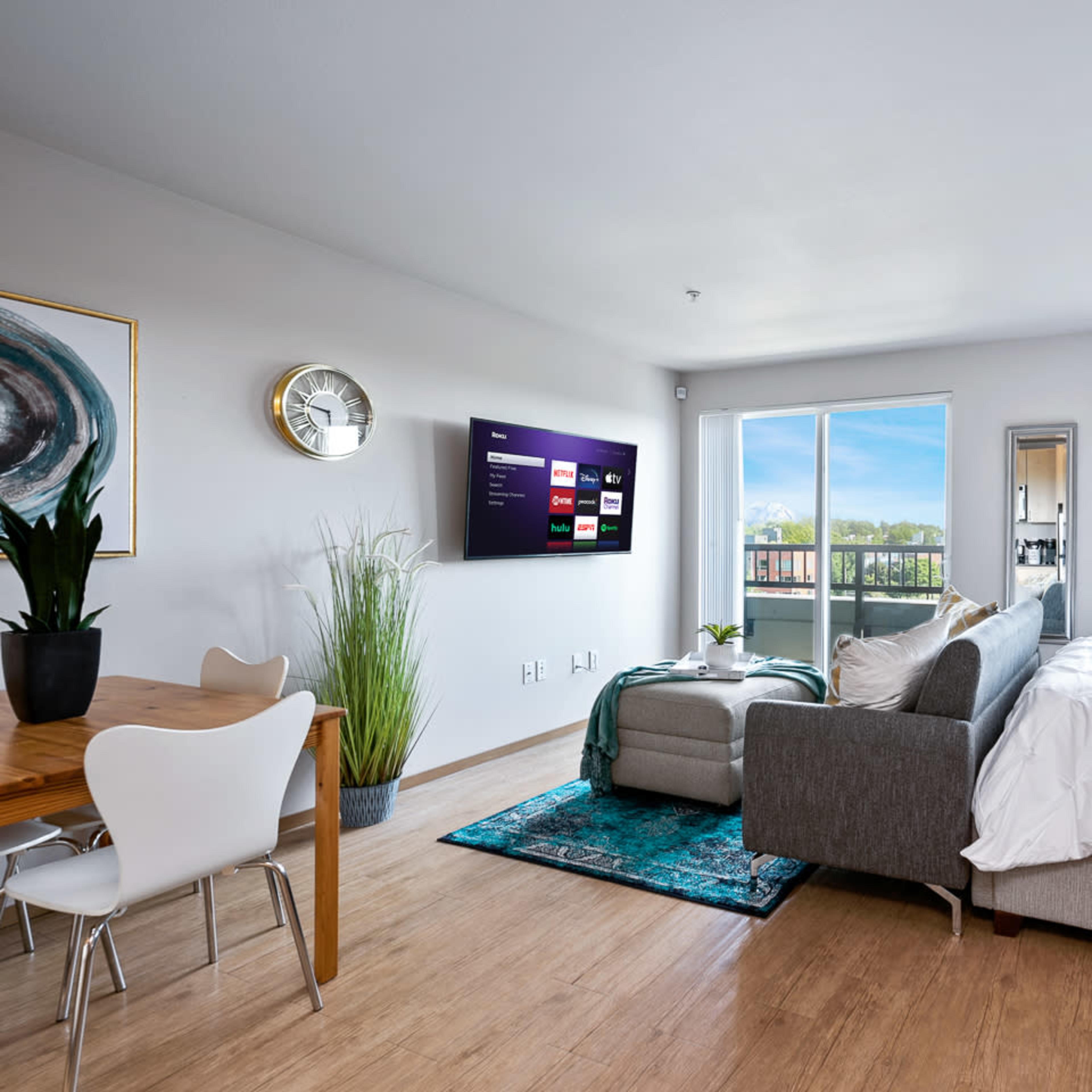 A modern studio apartment features a bed, a sofa, a dining table, and a wall-mounted TV with large windows displaying an outdoor view.