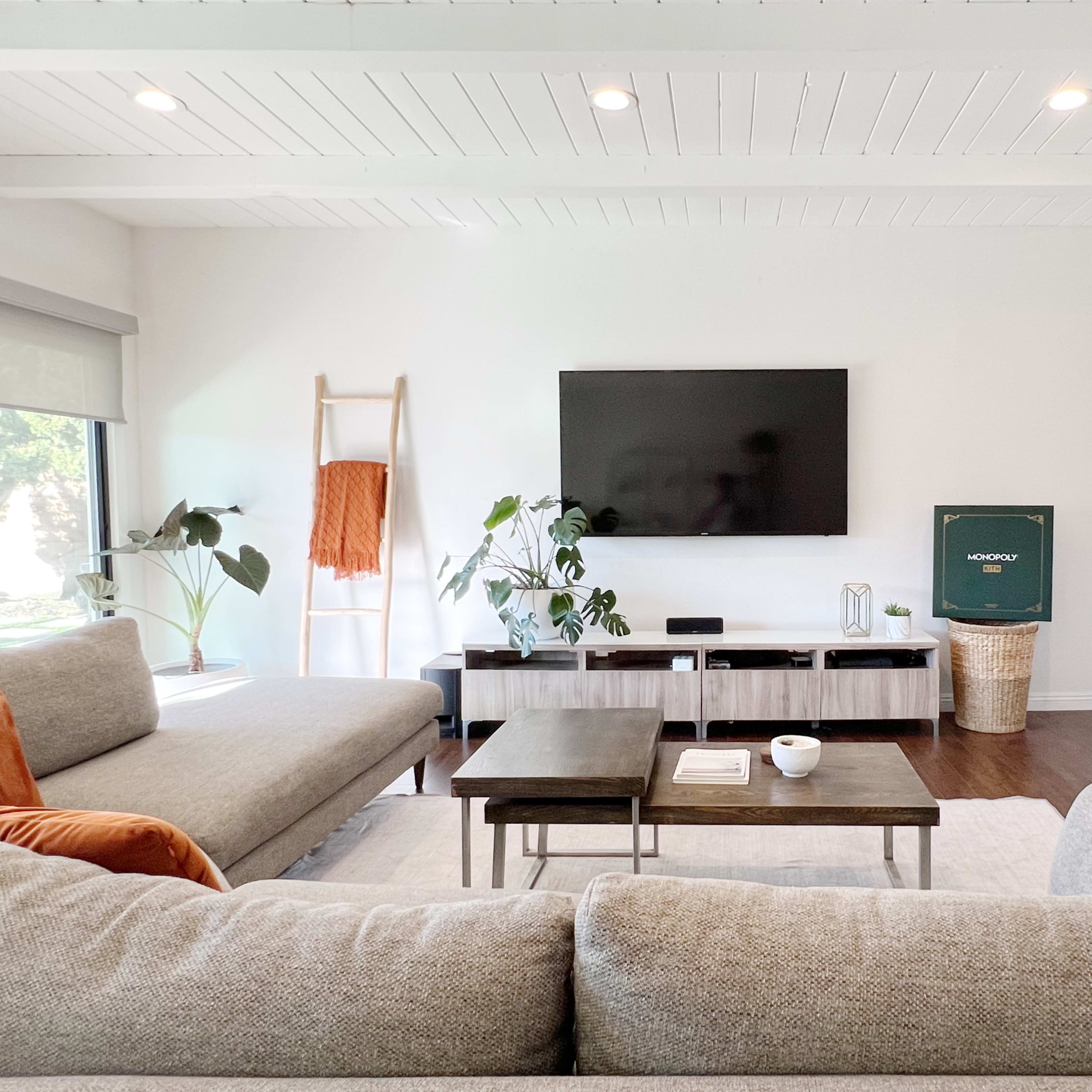 A modern living room features a gray sectional sofa, two coffee tables, a large TV mounted on the wall, and a sliding door leading to a bright space filled with plants.